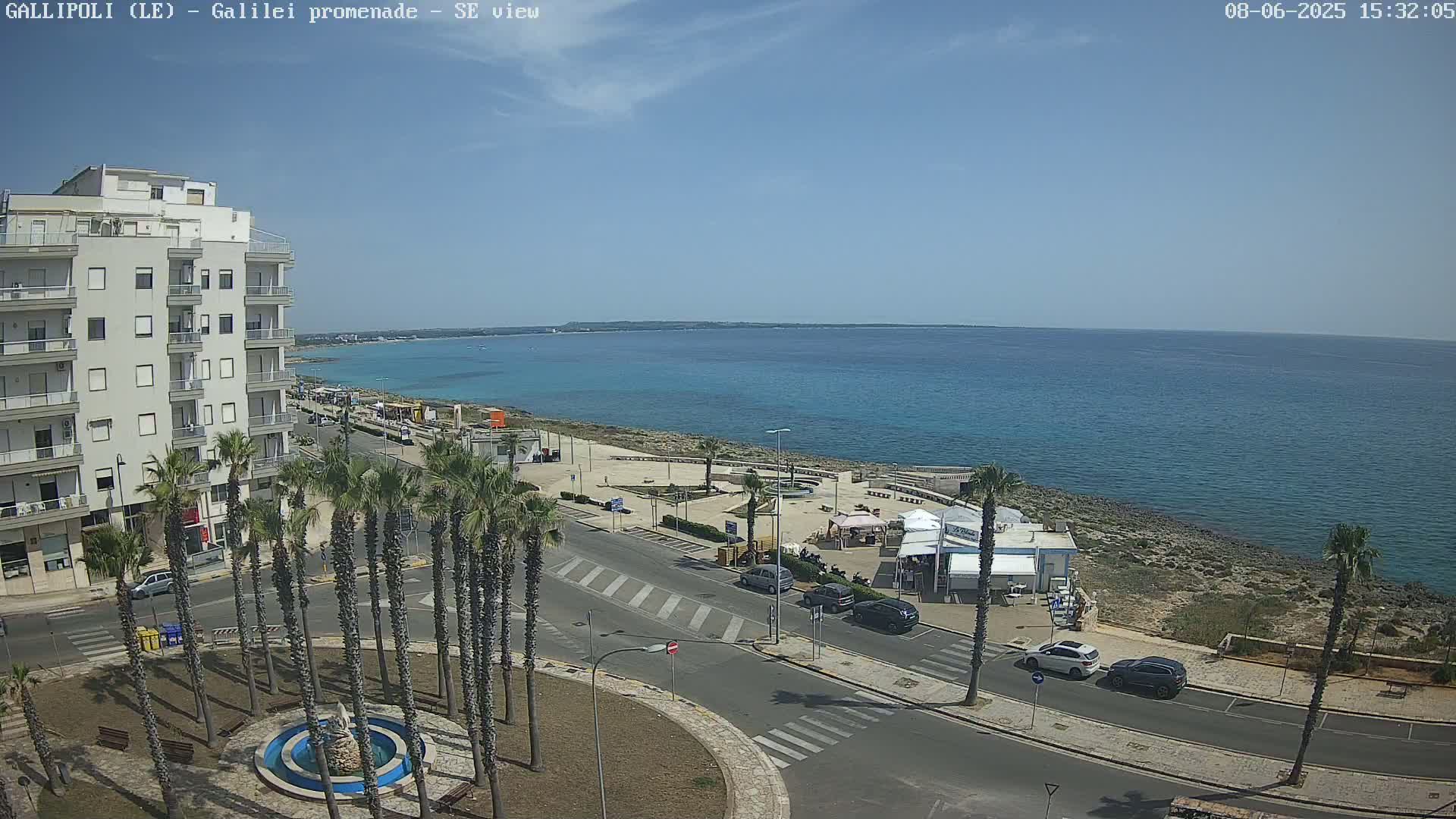 A seaside town scene on a sunny day shows a multi-story building, palm trees lining a street, parked cars, and a shoreline with clear blue water.