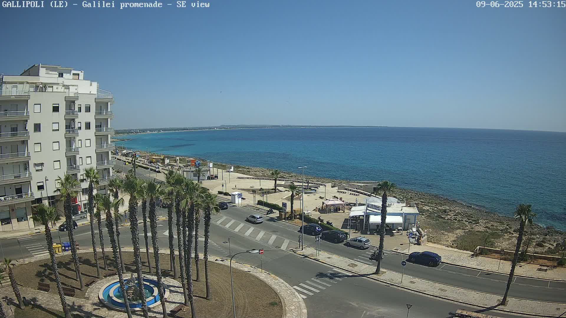 A coastal city scene on a sunny day shows a multi-story building, palm trees lining a street, a small beach area, and a calm blue sea extending to the horizon.