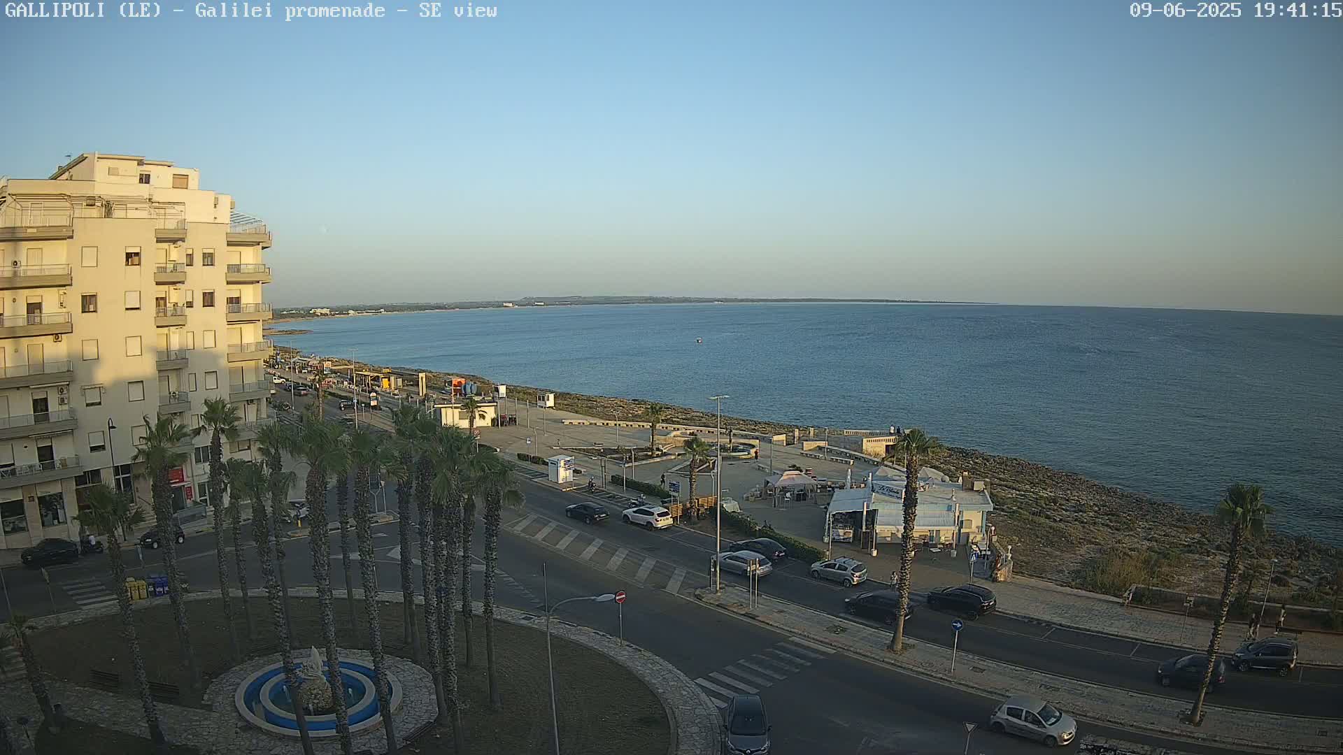 A seaside town at dusk, with calm water, a palm tree-lined road, and a multi-story building in the foreground.
