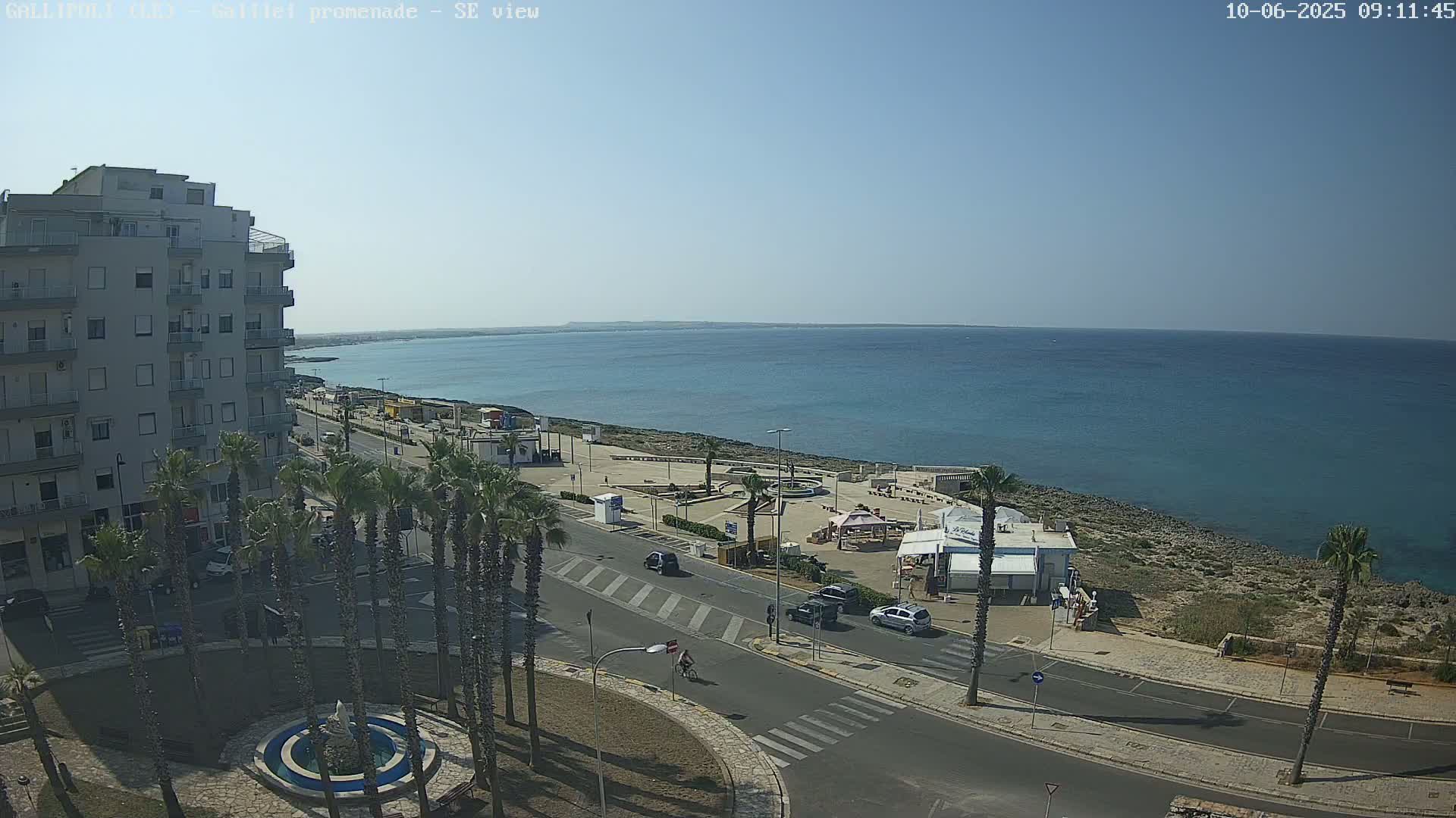 A seaside town on a sunny day shows a street lined with palm trees leading to a waterfront promenade with a few cars and people, and a building overlooking the scene.