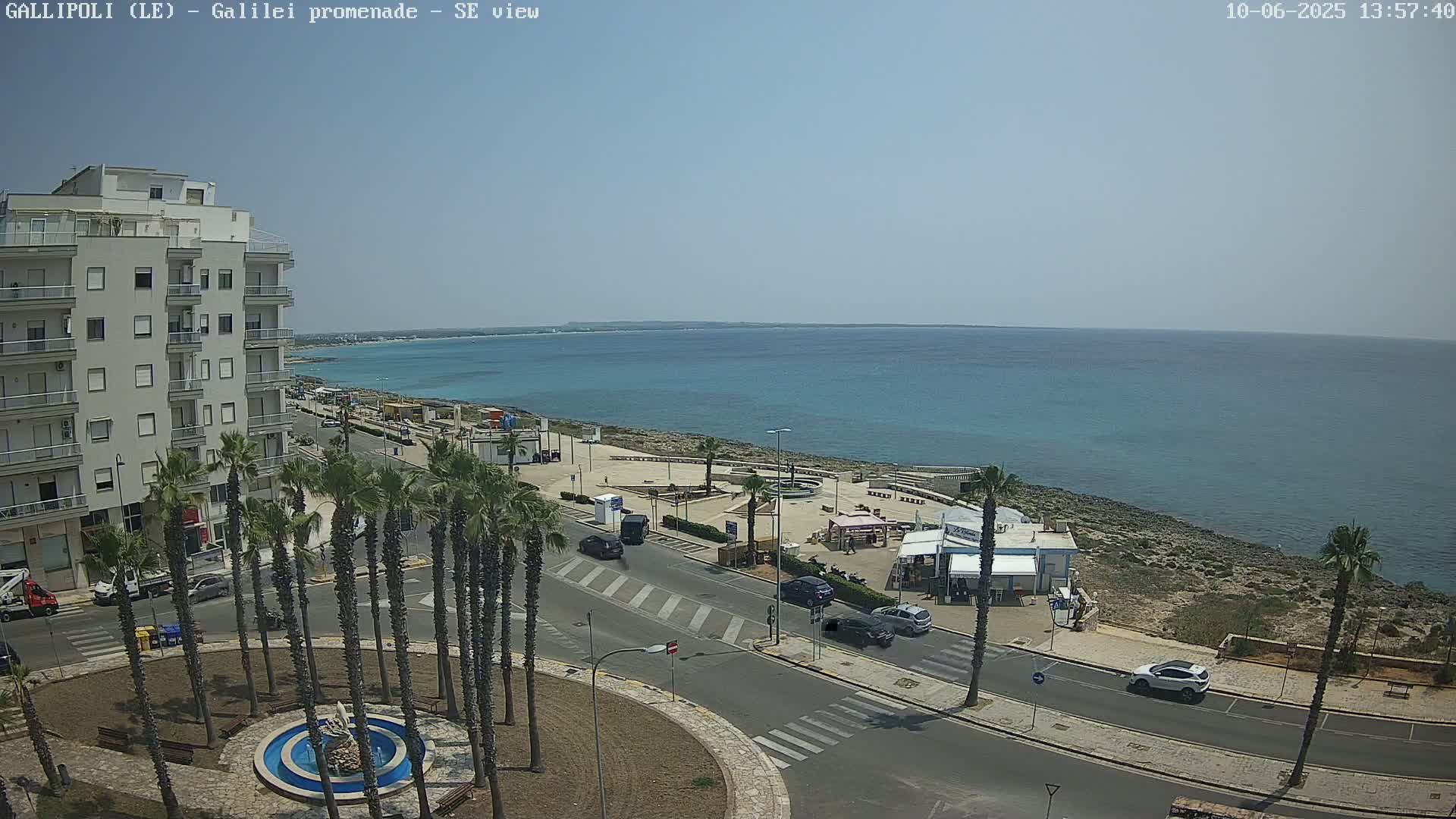 A seaside town on a sunny day shows a multi-story building, palm trees lining a roadway near a promenade overlooking a calm blue sea.