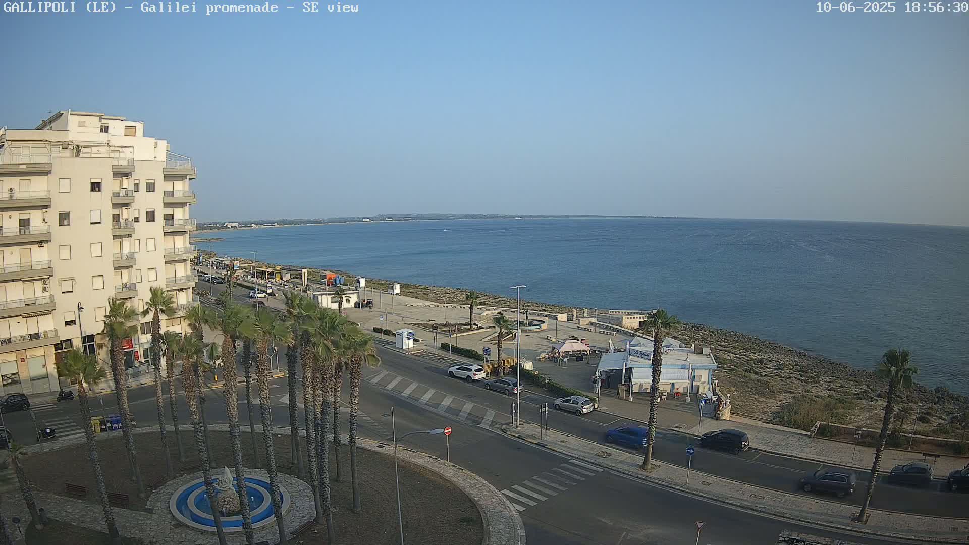 A seaside town scene on a clear, sunny day shows a multi-story building, palm trees lining a road leading to a promenade by a calm ocean.