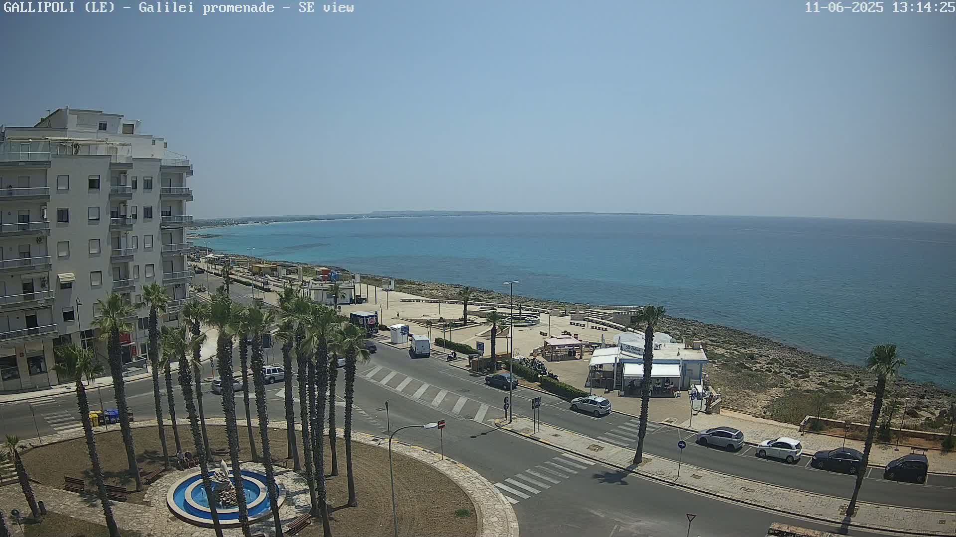 A seaside town on a sunny day, with a multi-story building, palm trees, and parked cars near a waterfront promenade overlooking a calm, turquoise sea.
