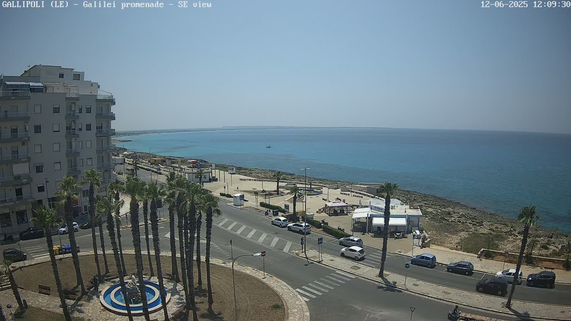 A sunny seaside town scene shows a multi-story building, a palm tree-lined roadway, parked cars, a small waterfront plaza, and a calm turquoise sea under a clear blue sky.