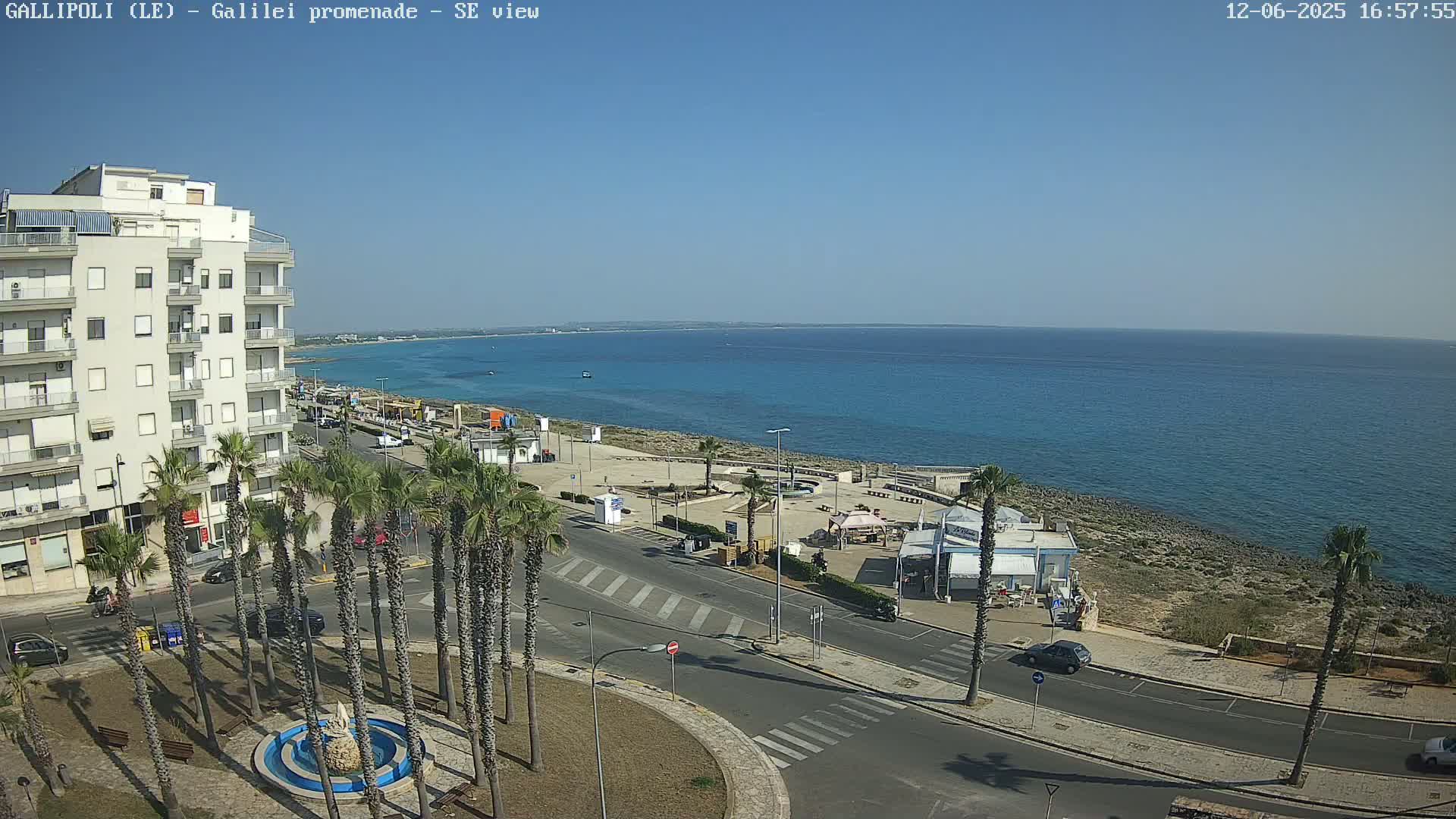 A clear, sunny day overlooks a coastal town with a multi-story building, palm trees, roads, and a promenade leading to a calm, blue sea.