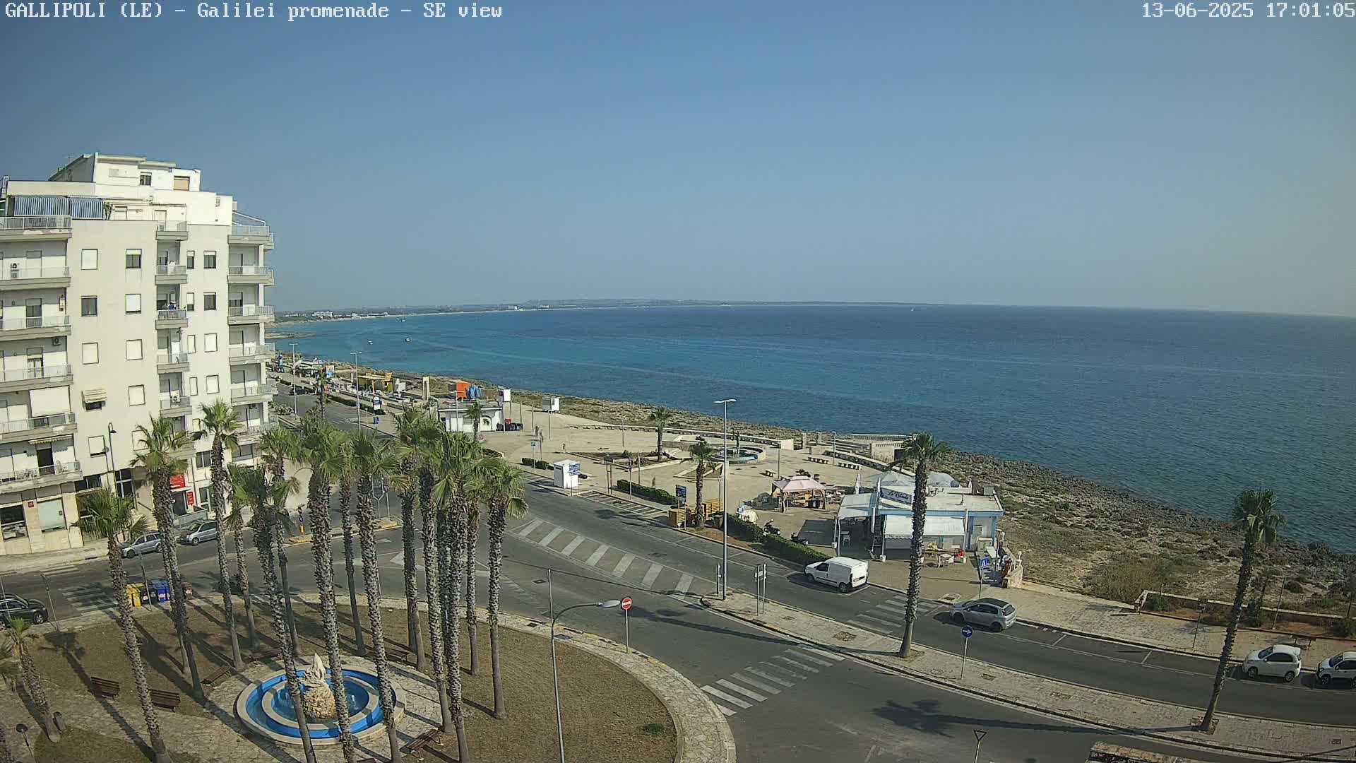 A seaside town scene on a sunny day shows a multi-story building, palm trees lining a street, a promenade, and calm blue water.