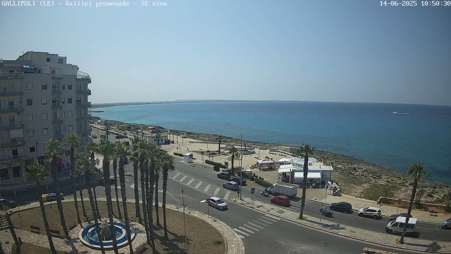 A sunny day overlooking a seaside town with a multi-story building, palm trees lining a road, a small plaza, and a clear blue ocean stretching to the horizon.