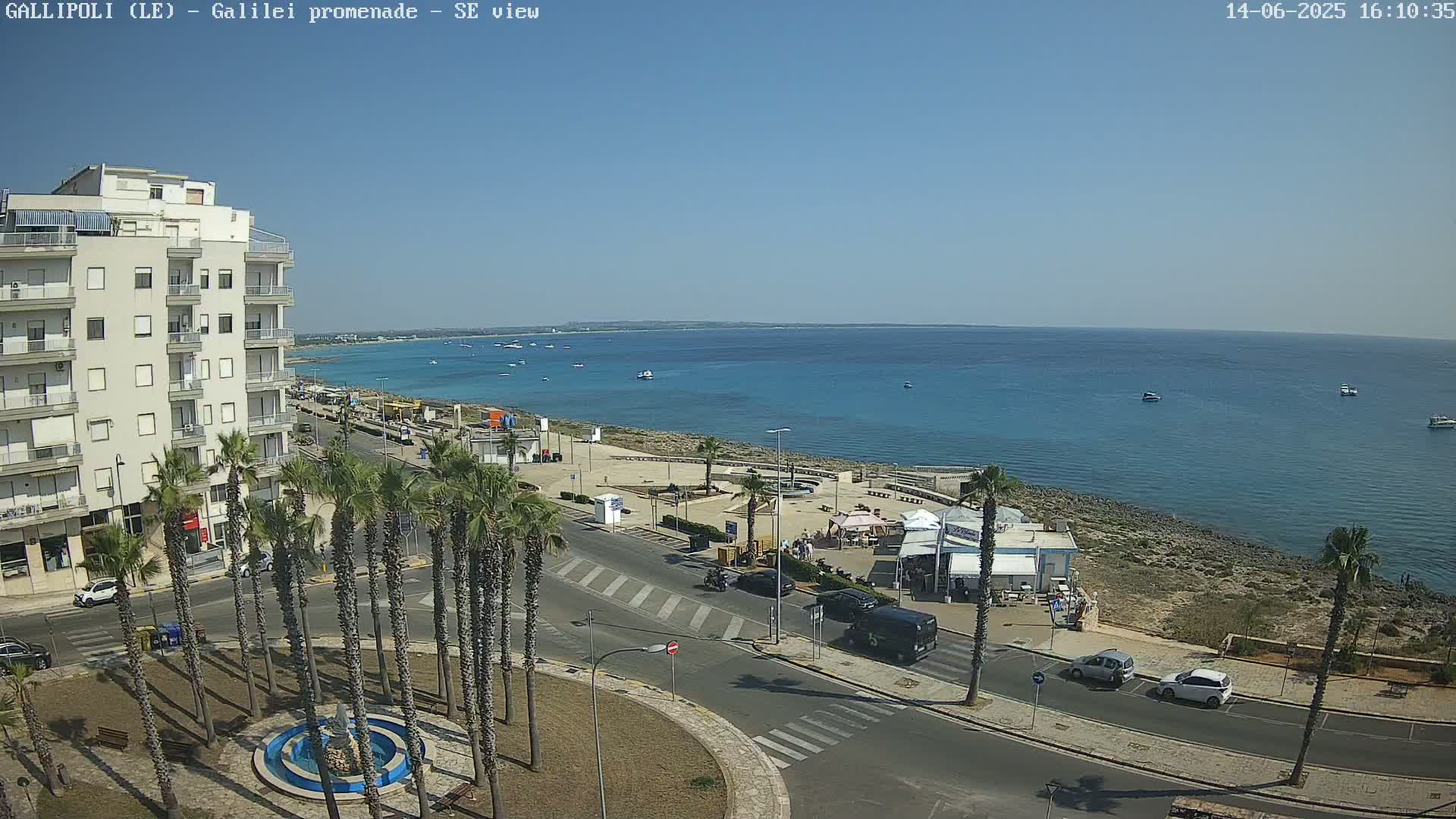 A seaside town scene on a sunny day shows a multi-story building, palm trees lining a roadway, a calm blue sea with boats, and a rocky shoreline with some outdoor seating areas.