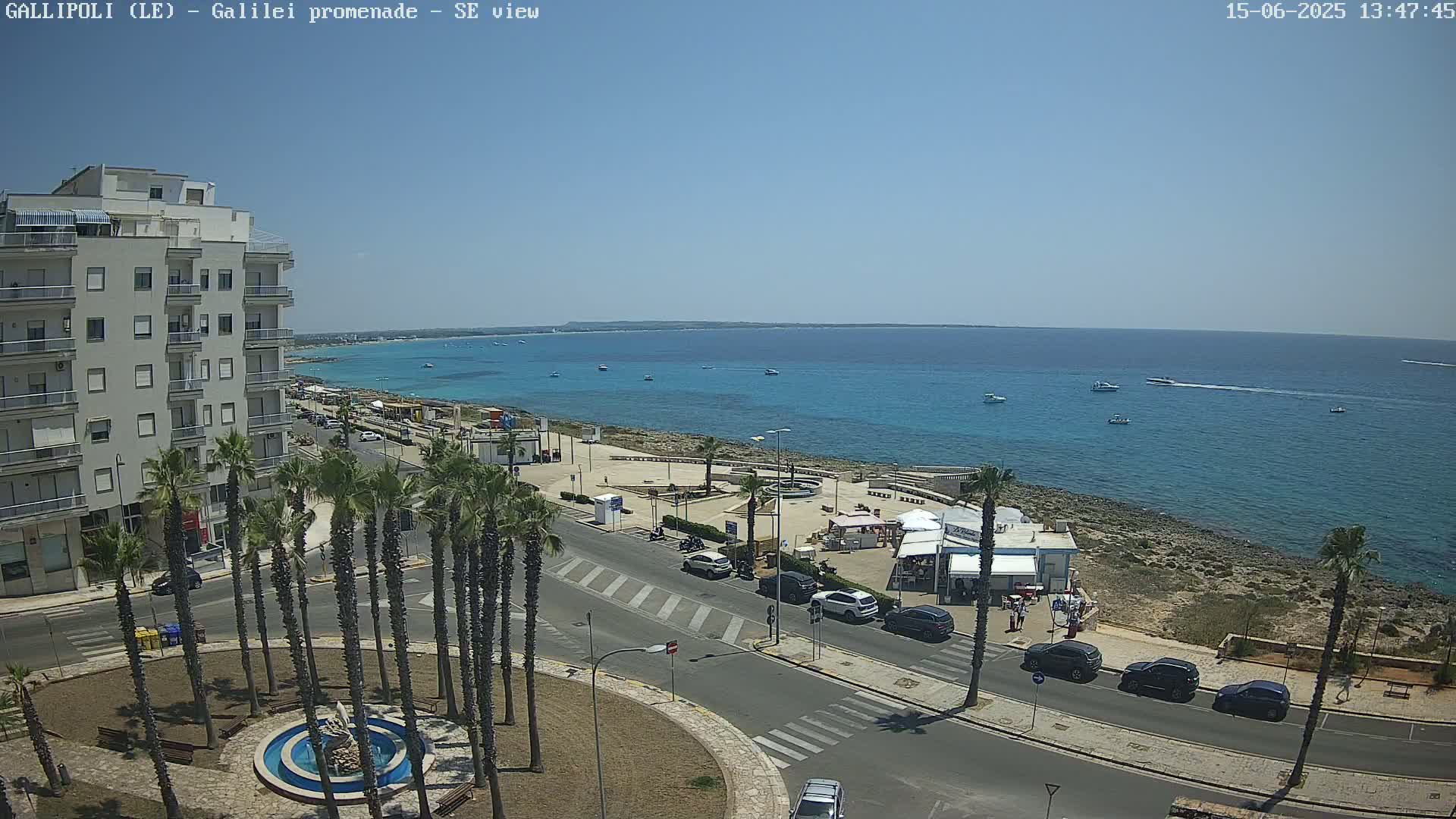 A sunny day reveals a coastal scene with a multi-story building, palm trees lining a road near a waterfront promenade, and several boats visible in the clear blue ocean.