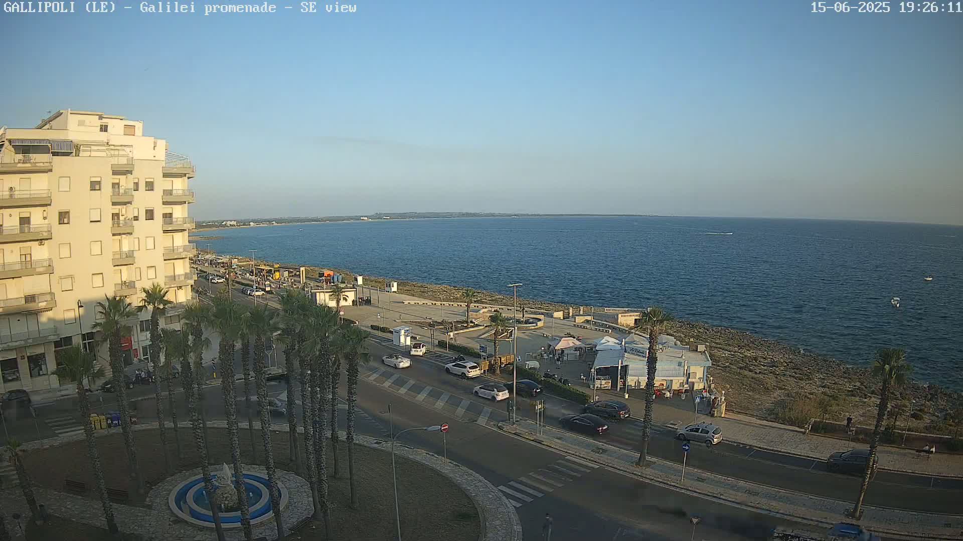 An elevated view of a seaside town shows a calm ocean under a clear blue sky, cars on a road lined with palm trees leading to a promenade with several small buildings.