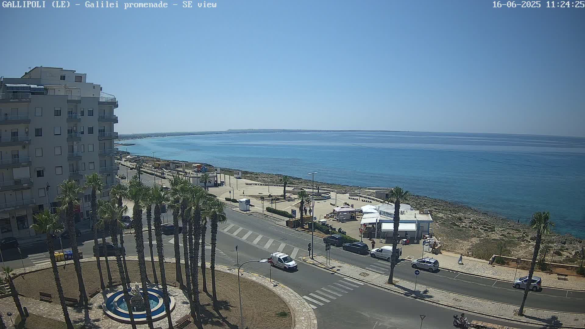 A high-angle view shows a sunny seaside town with palm trees, a roadway, and a calm blue ocean under a clear sky.