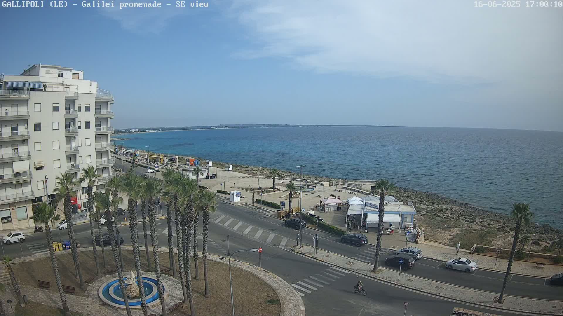 A seaside town scene on a sunny day shows a multi-story building, palm trees lining a roadway near a promenade, a calm ocean, and several cars parked along the street.