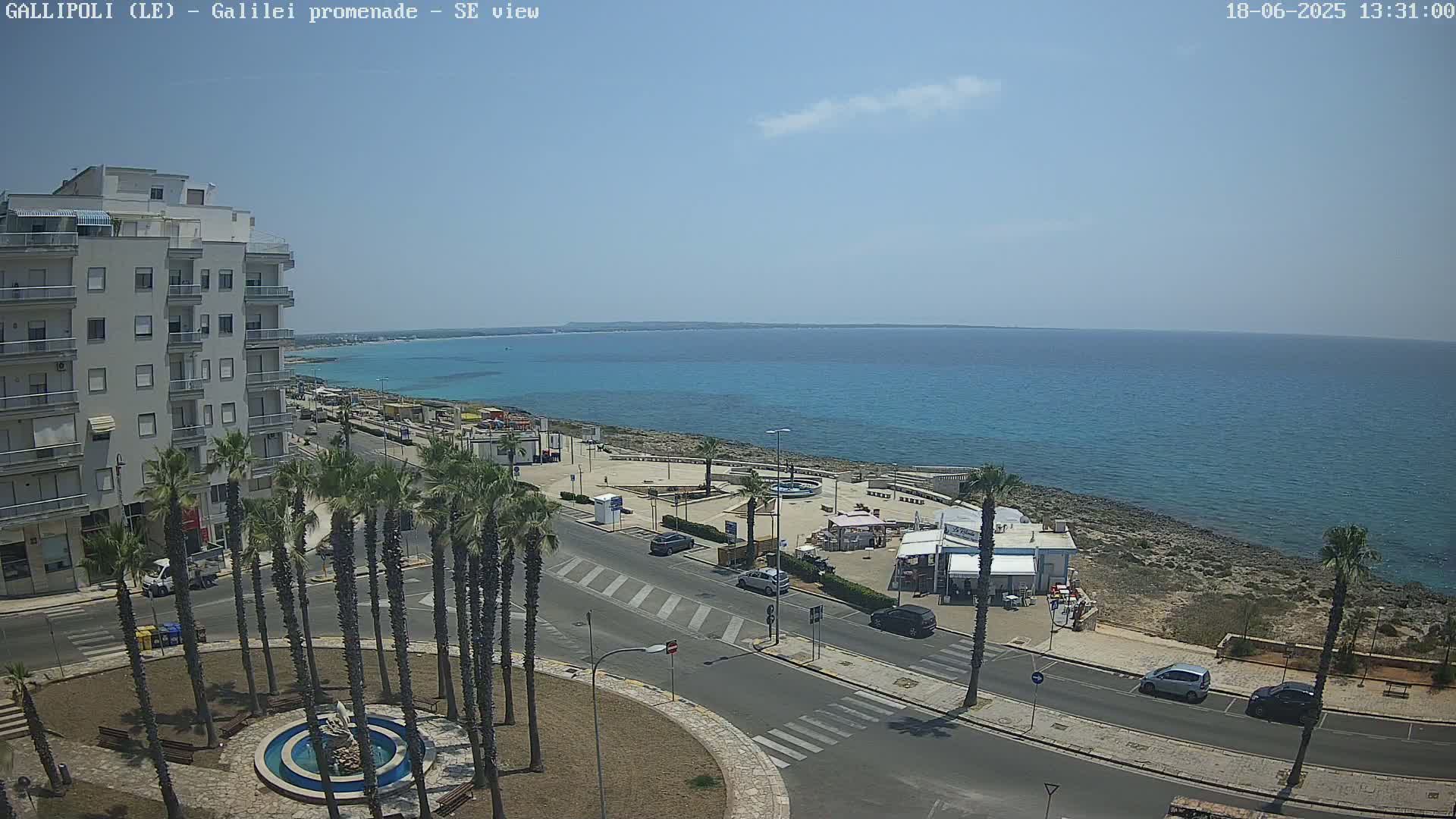 A seaside promenade with palm trees, a building, and several cars is visible on a sunny day, overlooking a calm turquoise sea.
