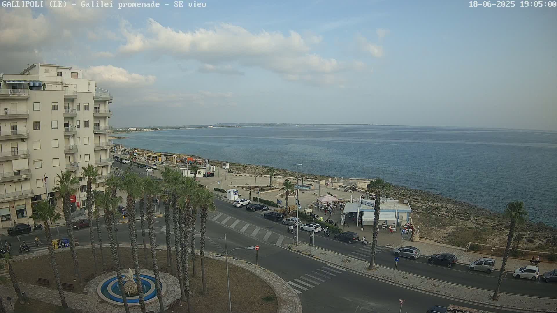 A partly cloudy day overlooks a coastal town with a seaside promenade, palm trees, cars parked along the road, and a fountain in a roundabout.