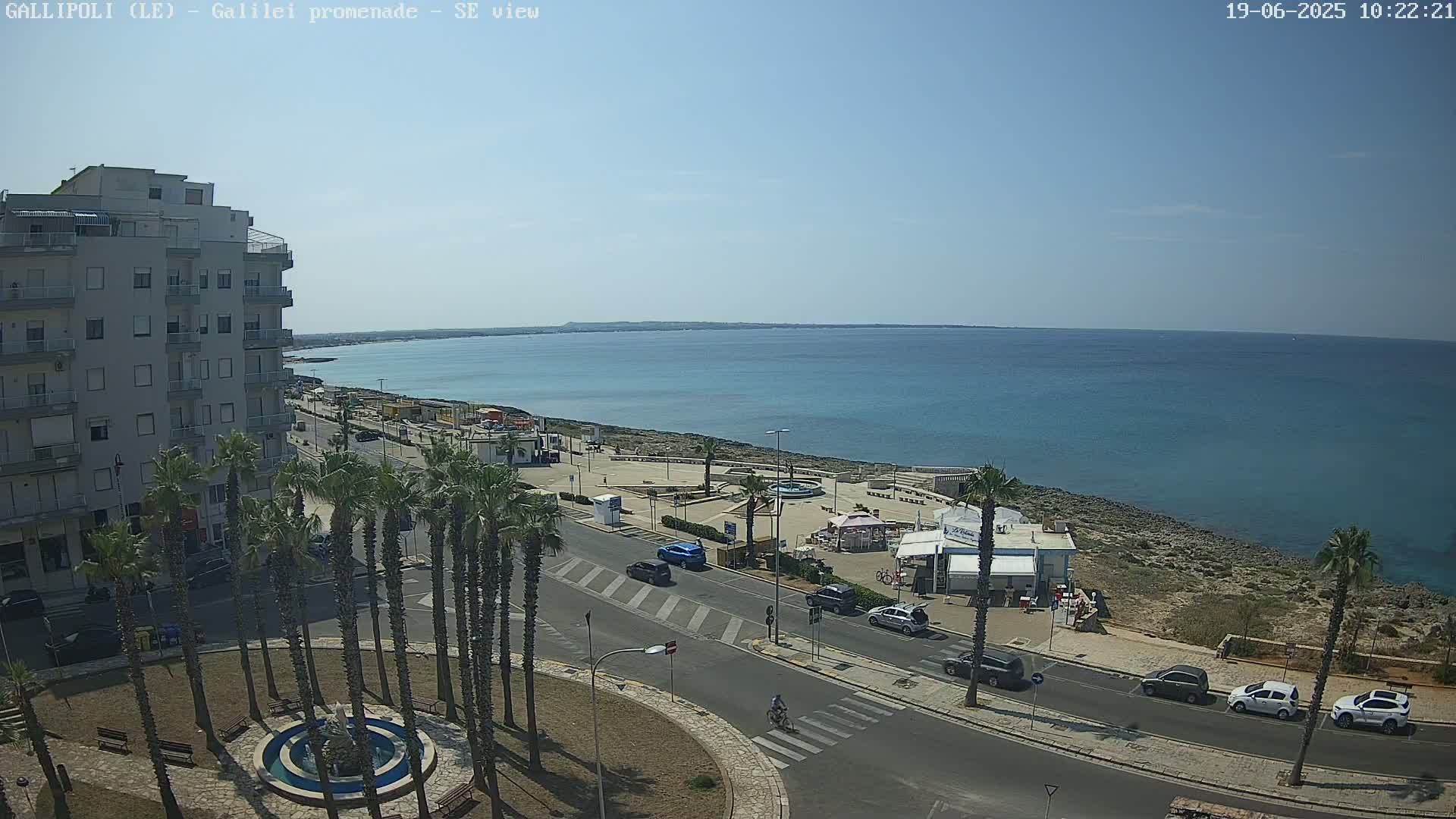 A seaside town scene on a sunny day shows a multi-story building, palm trees lining a street, cars parked along the road, and a calm blue sea in the background.