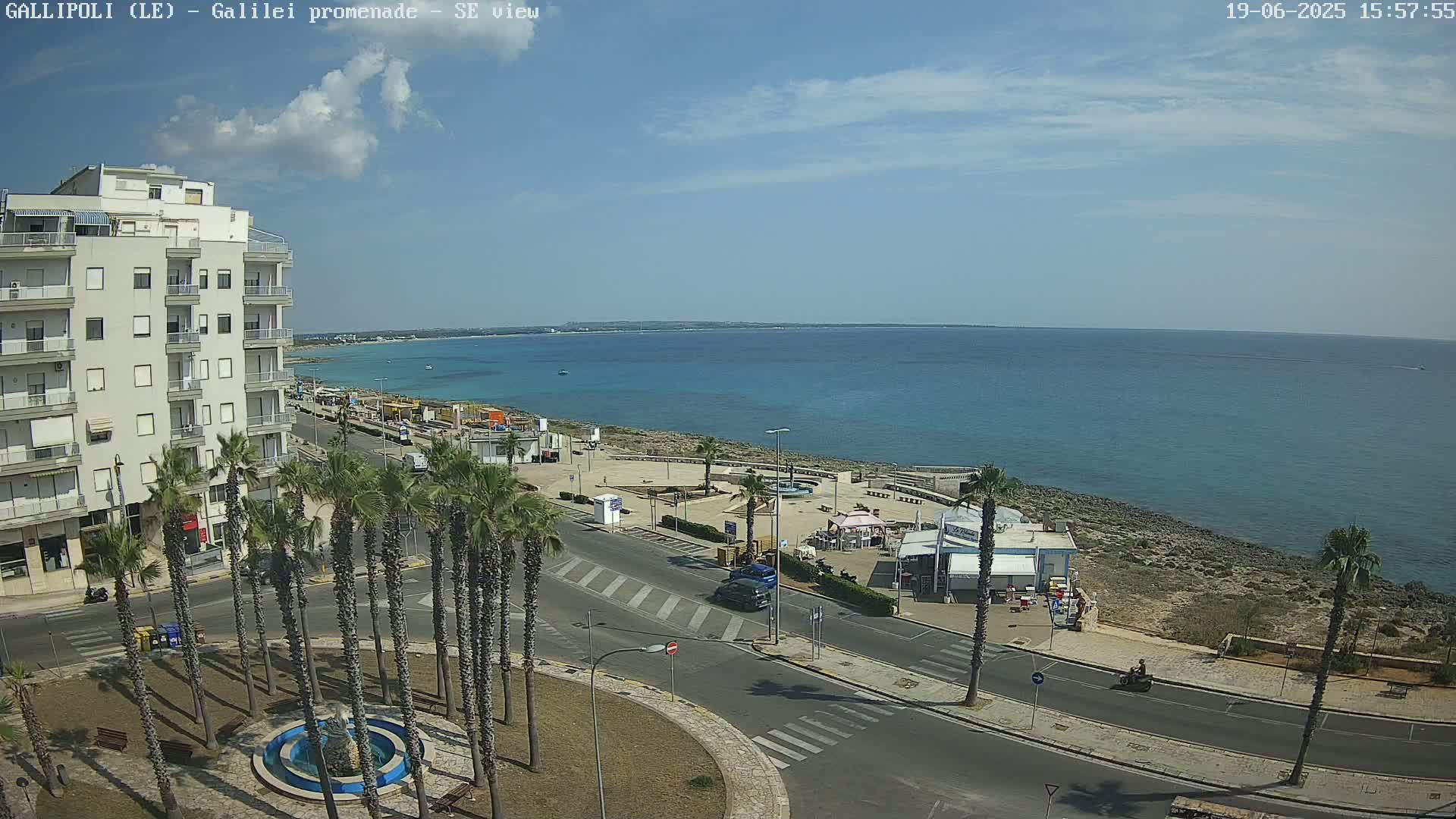 A partly cloudy day shows a seaside town with a multi-story building, palm trees lining a road leading to a beach with turquoise water.