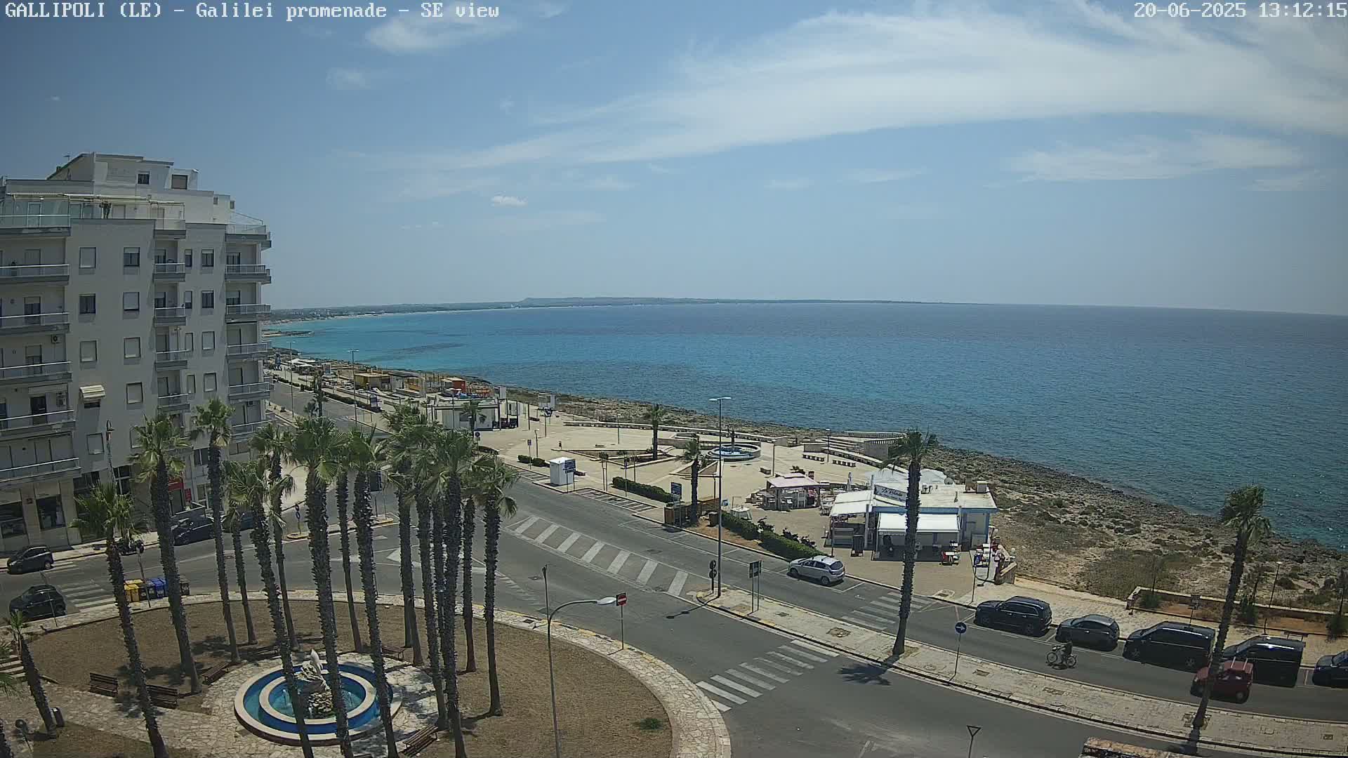 A sunny day shows a coastal town scene with a multi-story building, palm trees lining a roadway, a seaside promenade, and parked cars near turquoise water.