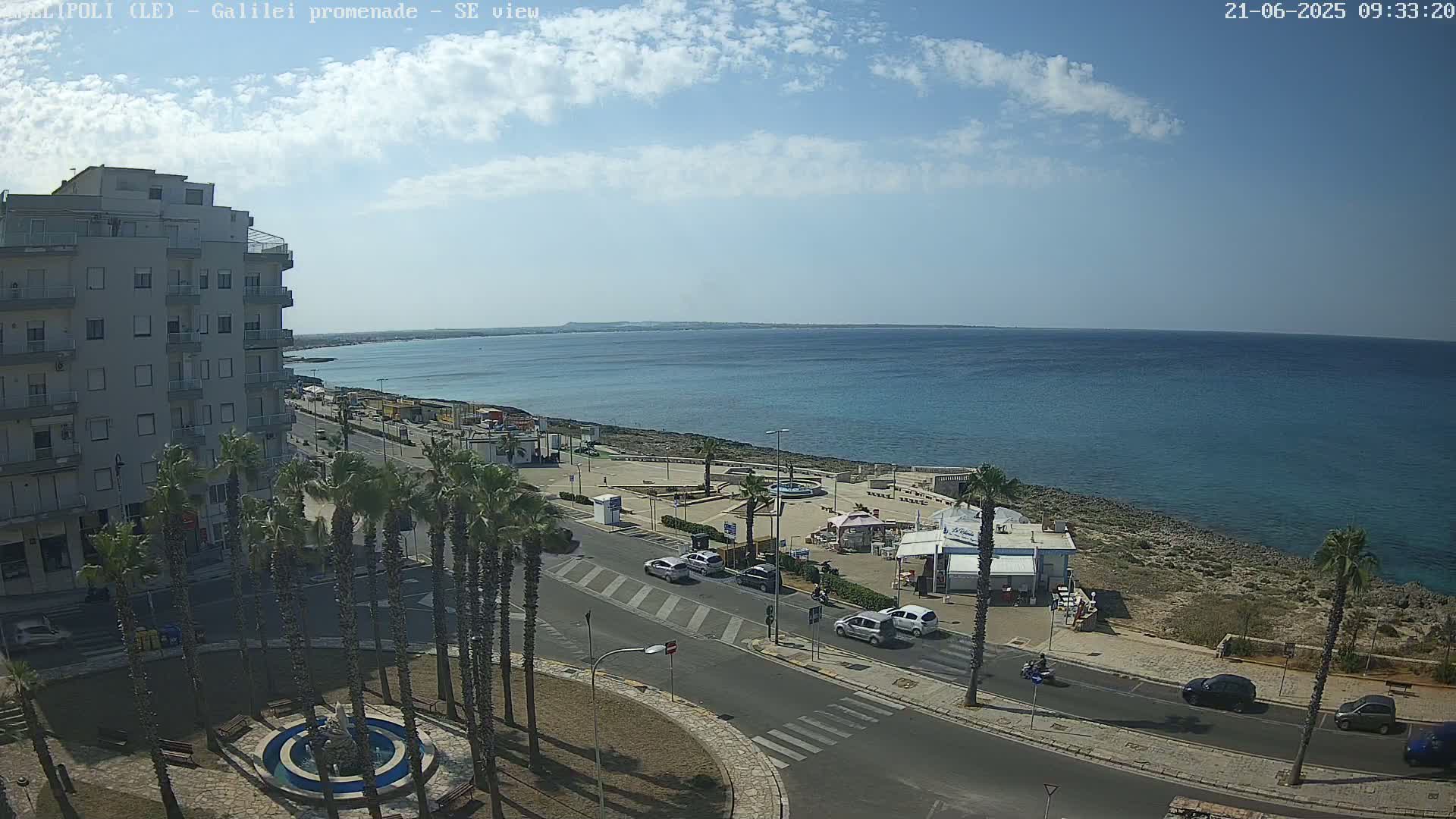 A seaside town scene under a partly cloudy sky shows a multi-story building, palm trees, cars parked along a road, and a small beachfront restaurant next to a calm blue sea.