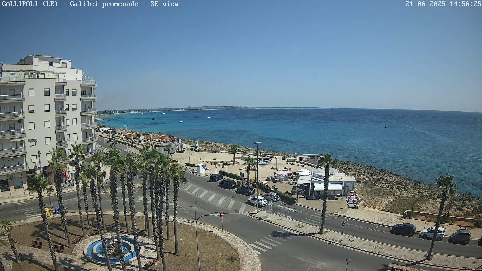 A seaside town scene on a sunny day shows a multi-story building, palm trees lining a street, a small plaza with a fountain, cars parked along the road, and a beach with turquoise water extending to a distant horizon.
