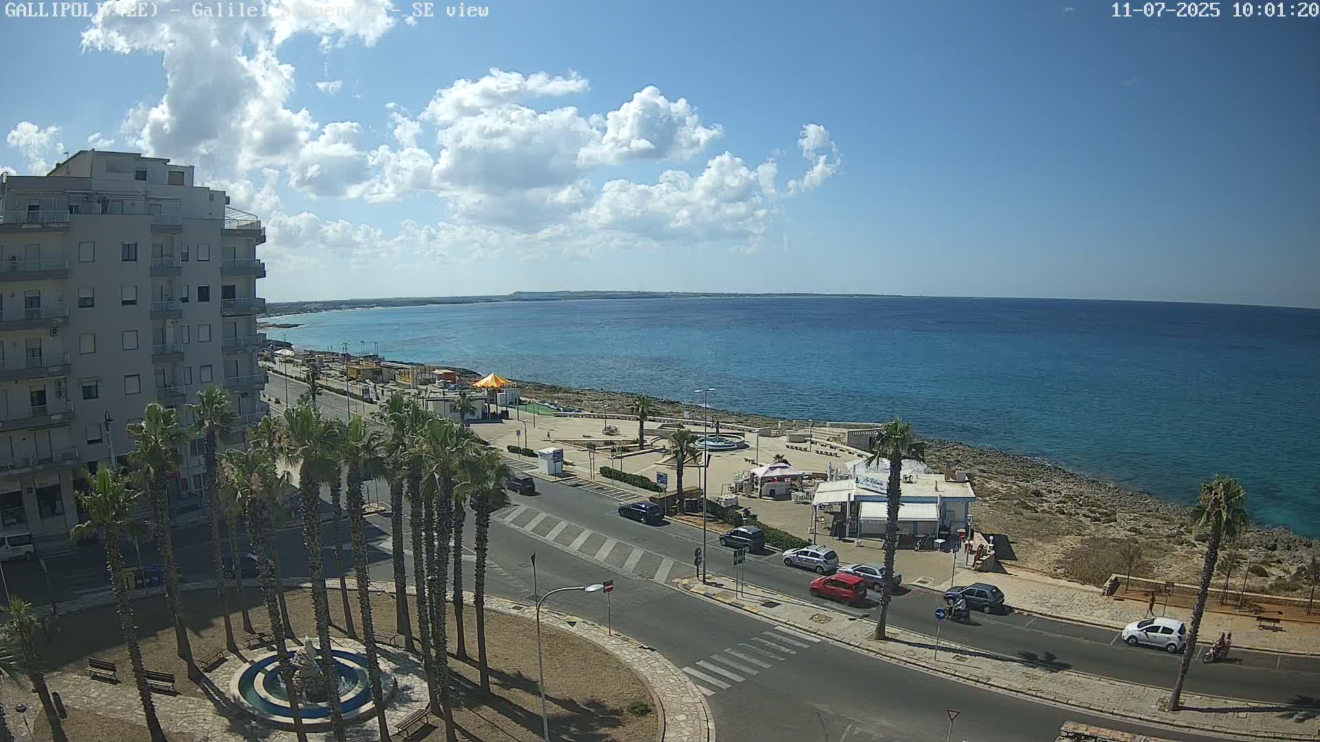 A partly cloudy sunny day shows a coastal town scene with a multi-story building, palm trees, a road, and a beachfront area with a few small buildings and calm blue water.