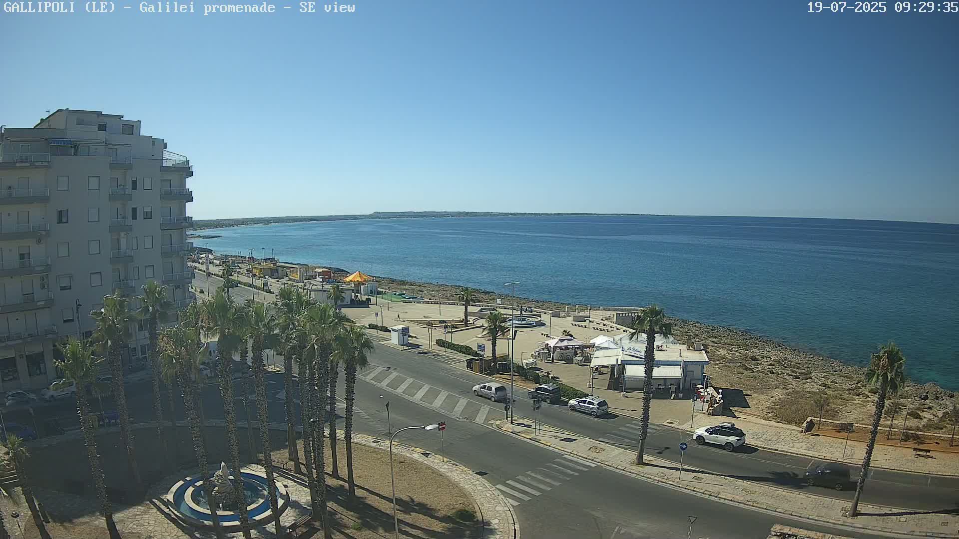 A seaside town on a sunny day shows a calm blue sea, a palm tree-lined promenade, a multi-story building, and several cars parked and driving along the street.
