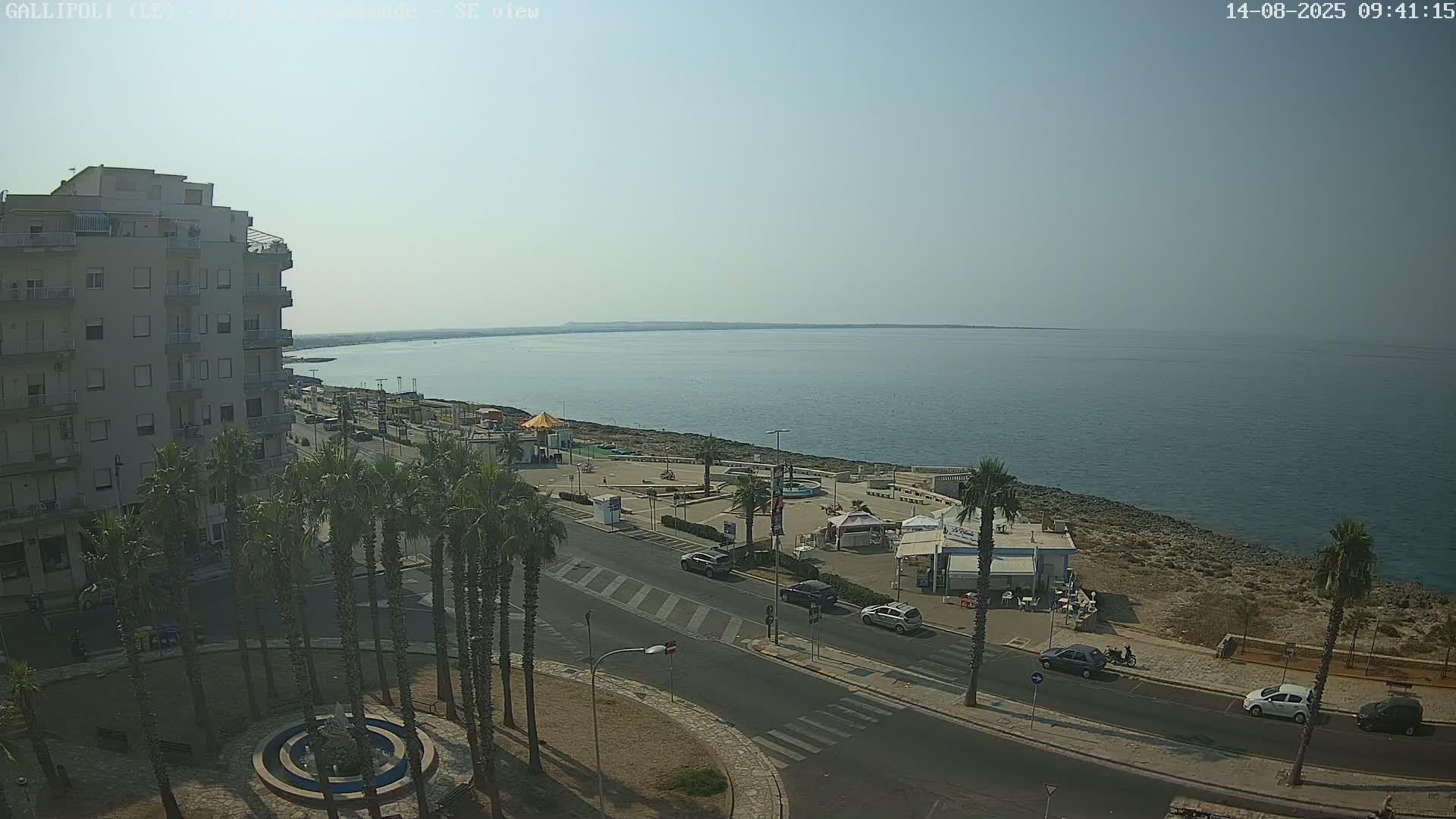 A coastal town scene on a sunny day shows a multi-story building, palm trees lining a street, and a calm sea extending to the horizon.
