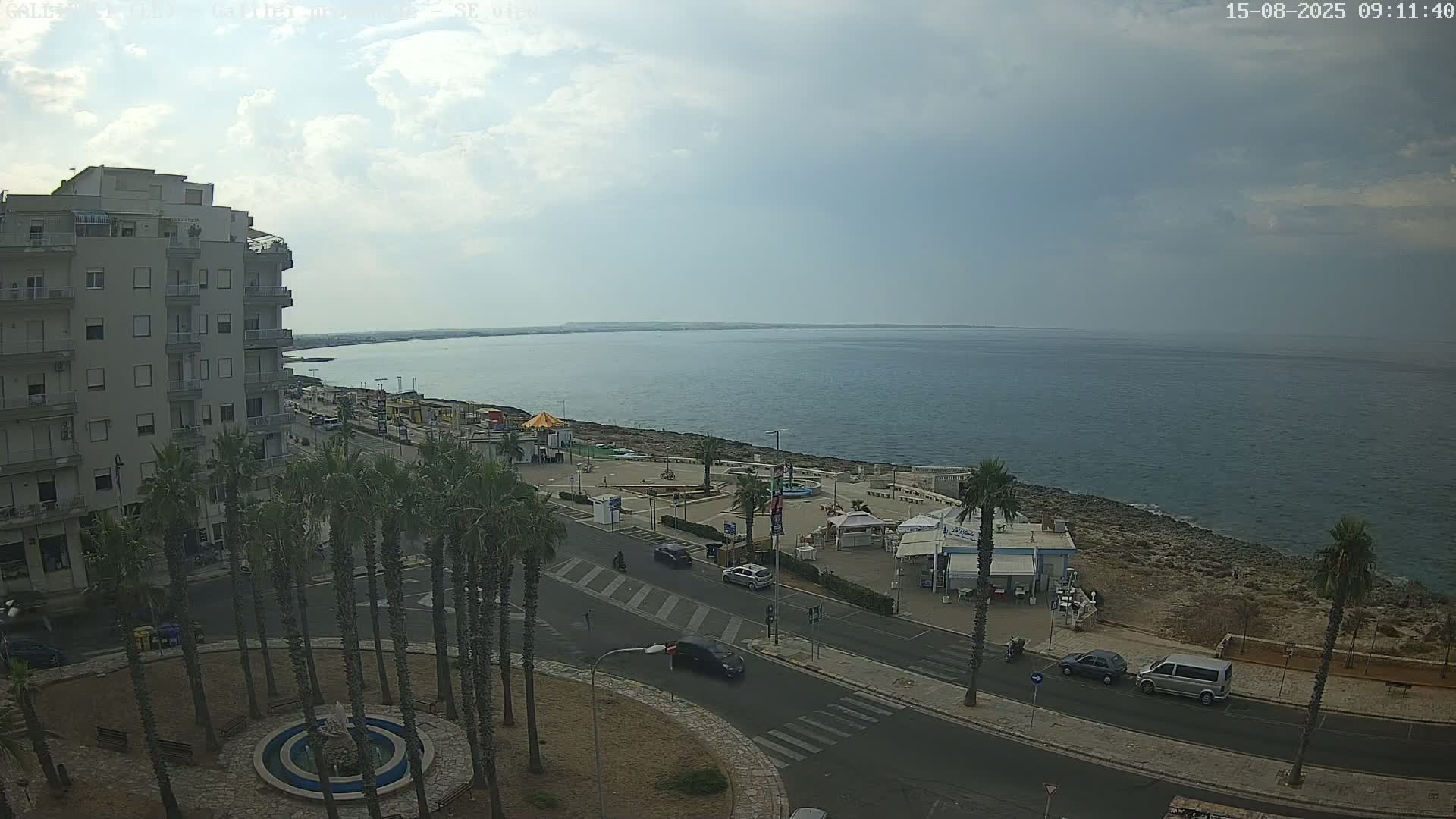 A partly cloudy day reveals a coastal town scene with palm trees lining a street next to a calm sea, and a multi-story building visible in the background.