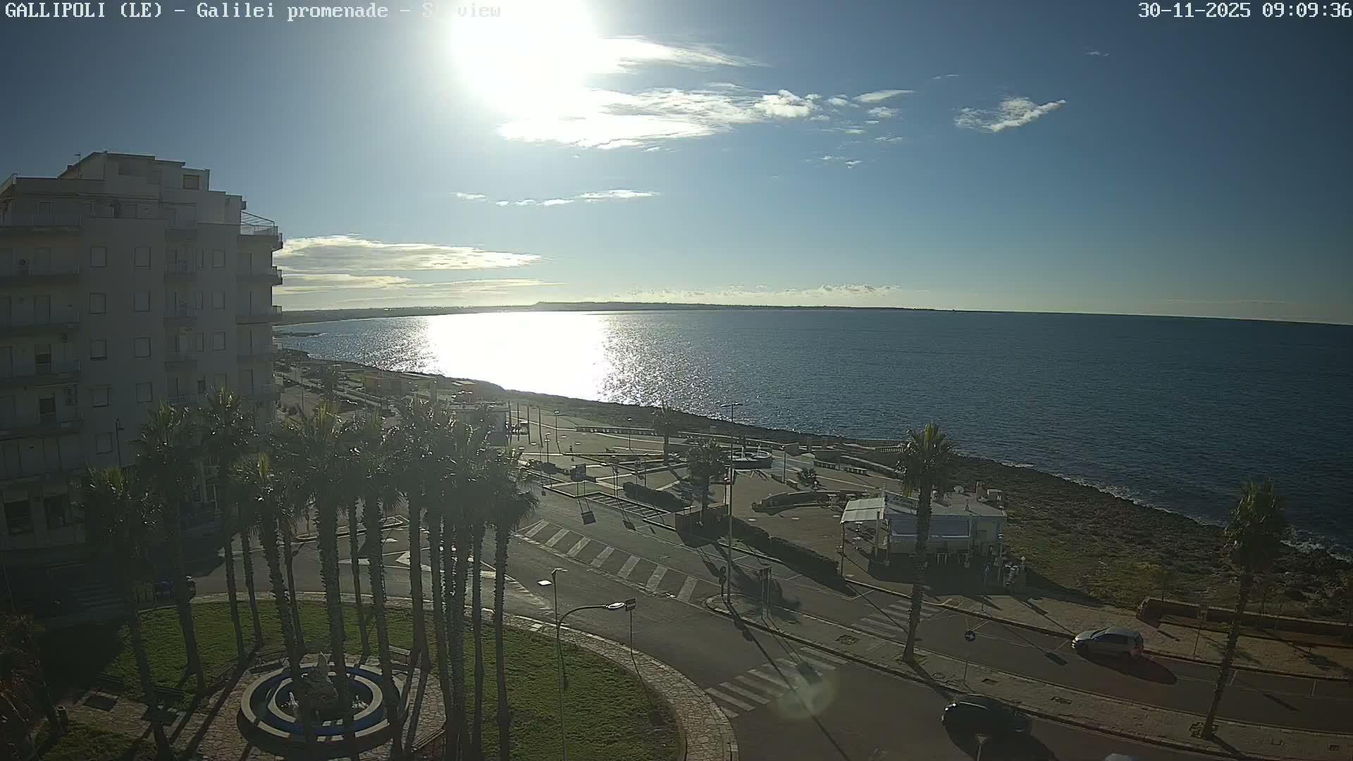 On a bright, sunny day with a clear blue sky and scattered white clouds, a coastal scene is visible with a calm, sun-drenched sea, a promenade lined with palm trees and a circular garden, and a tall building to the left.