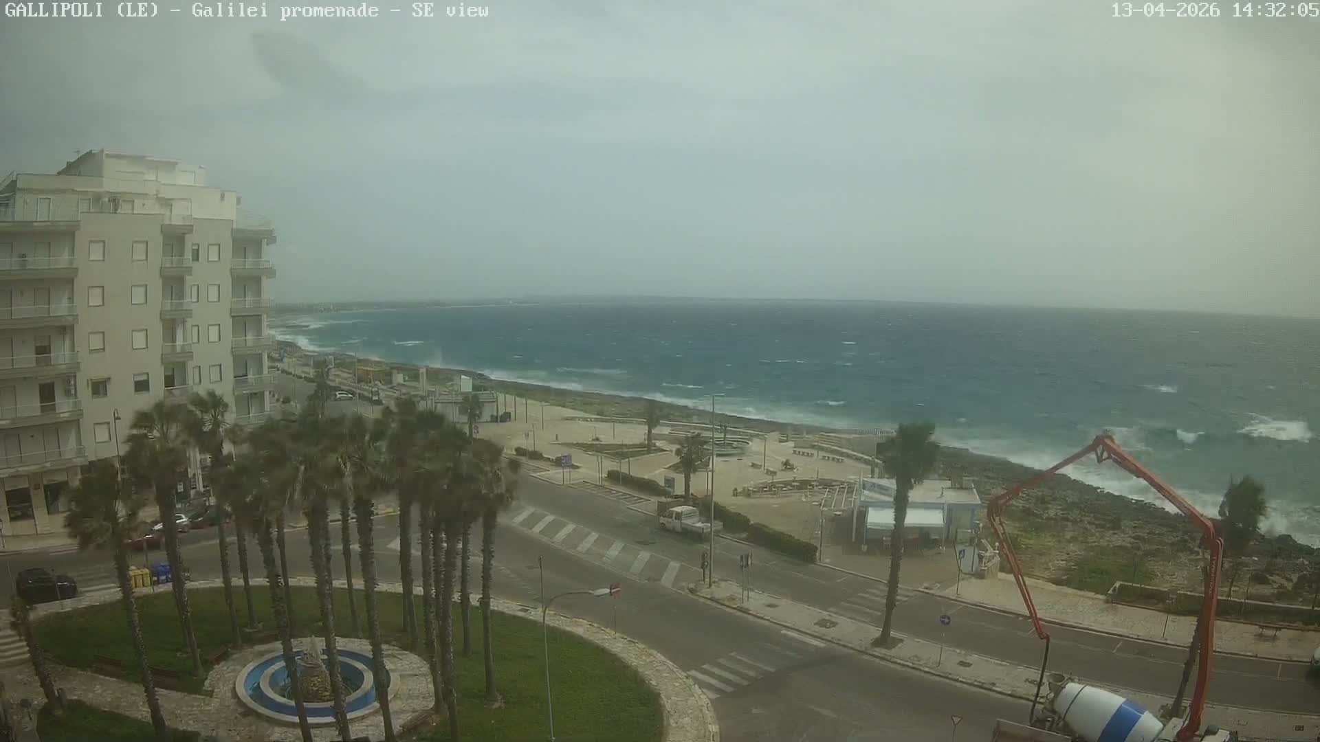 A stormy coastal scene depicts a multi-story building overlooking a wet promenade lined with palm trees, a road, and a turbulent grey-blue sea with crashing waves under an overcast sky.