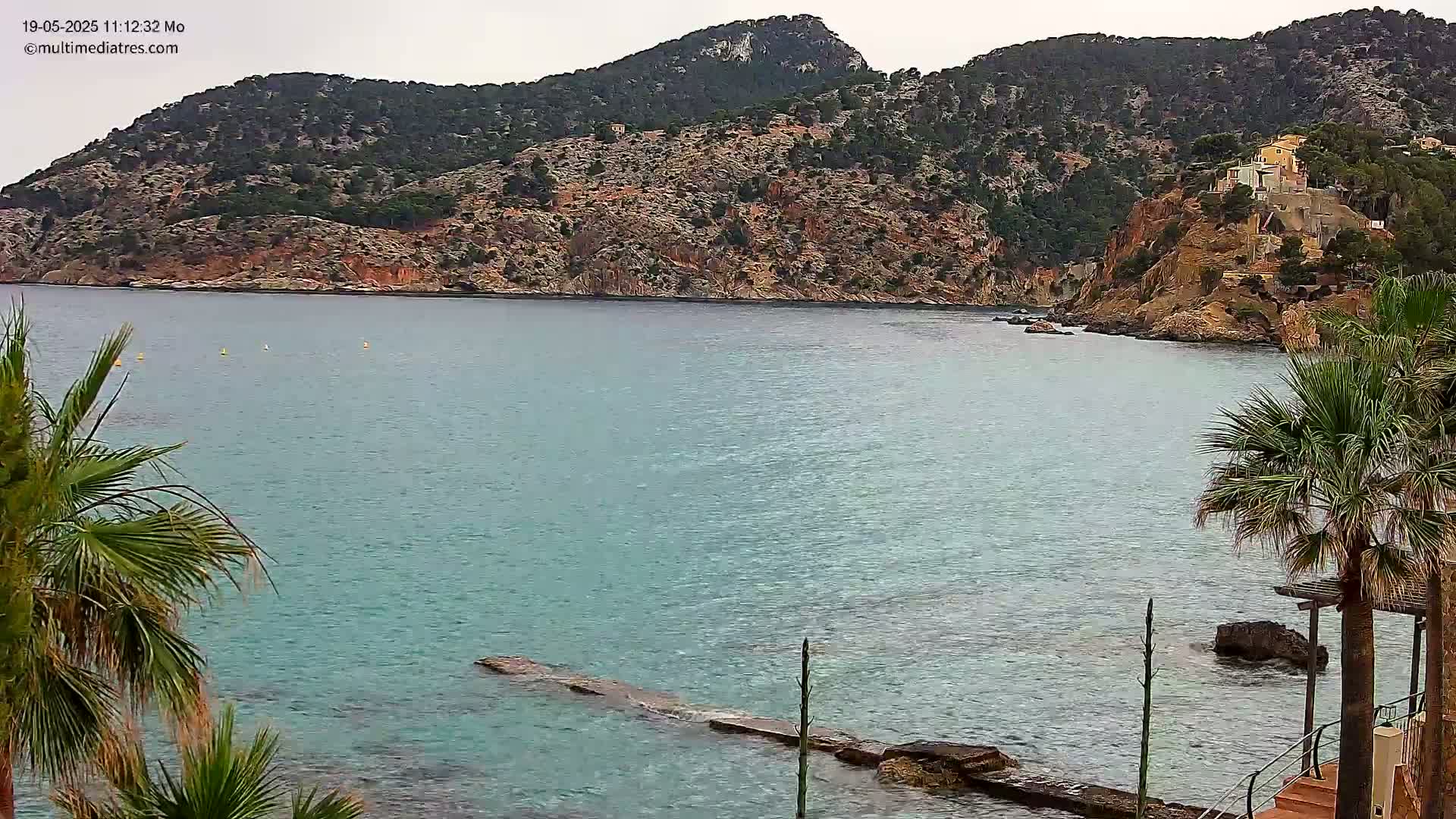 A calm, turquoise bay is nestled between rocky hillsides dotted with vegetation and a few buildings, under an overcast sky.