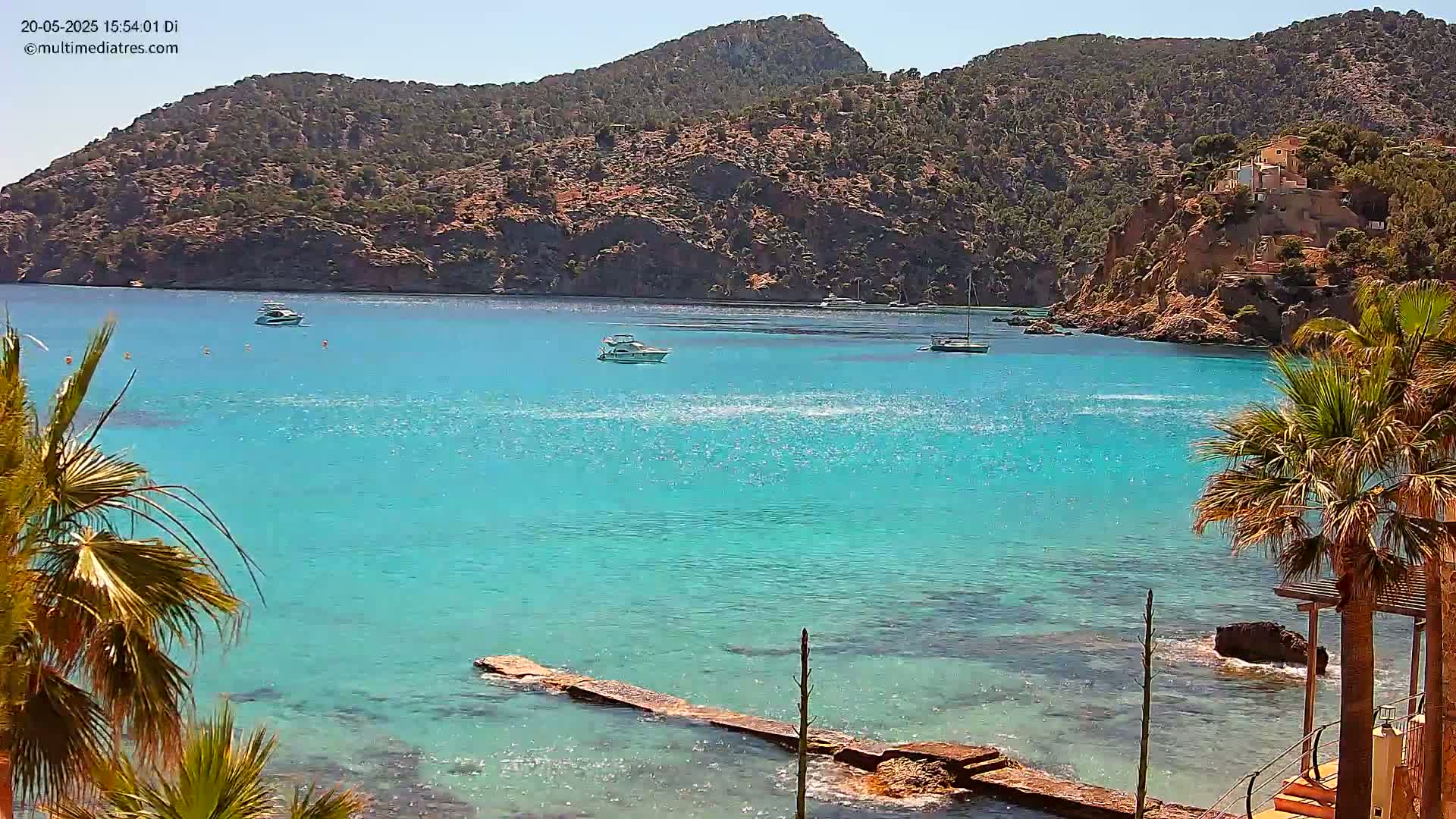 Bright turquoise water laps a rocky shore lined with palm trees, under a sunny sky, with several boats visible in the bay and a mountainous backdrop.