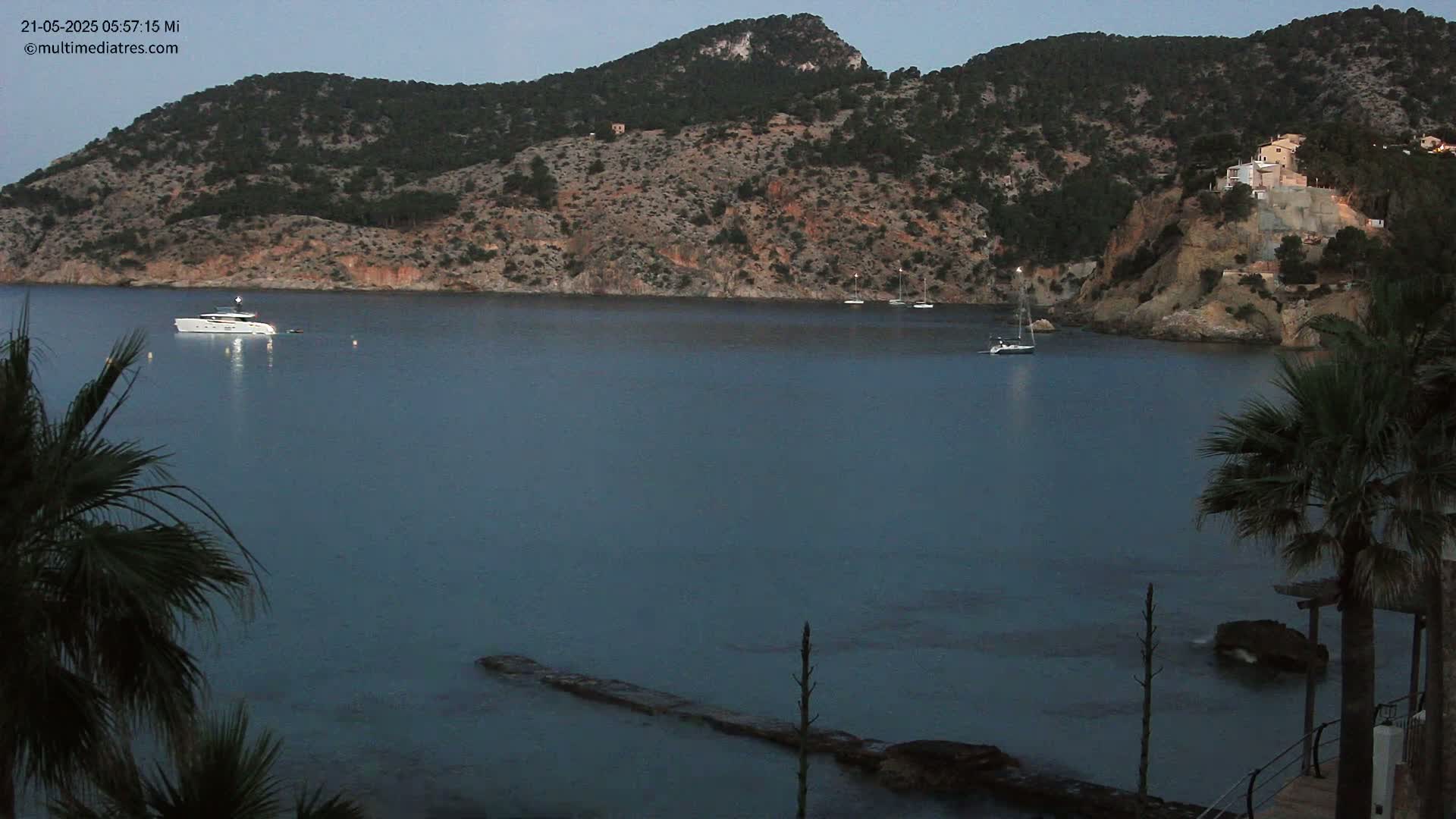 A calm, dark-blue bay at dusk is nestled between rocky hillsides dotted with sparse vegetation, with a few boats and a yacht visible on the water.