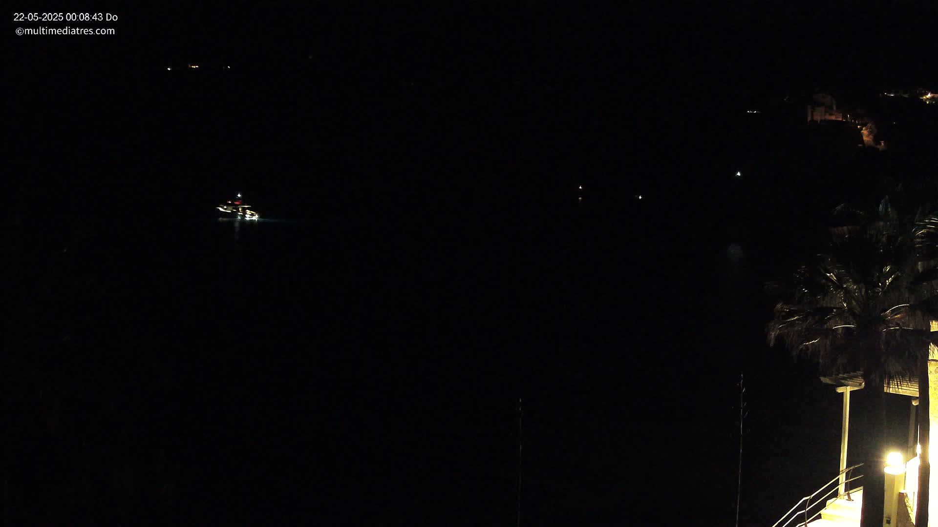 A dark nighttime scene shows a small boat with lights on the water, distant lights on the shore, and a palm tree partially visible in the lower right corner.