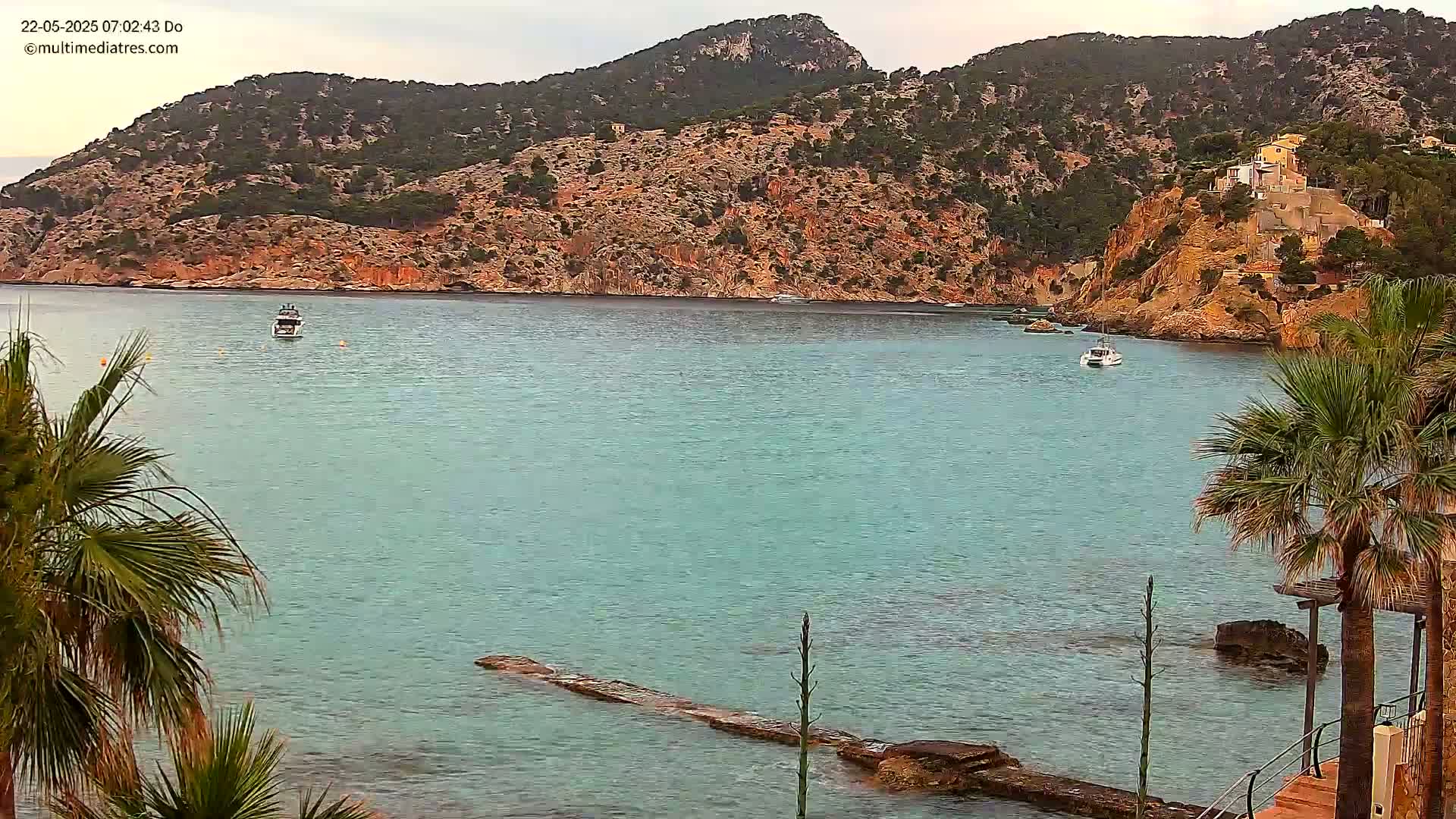 A calm, teal-colored bay is nestled between rocky hillsides dotted with Mediterranean vegetation, with a few boats visible on the water.