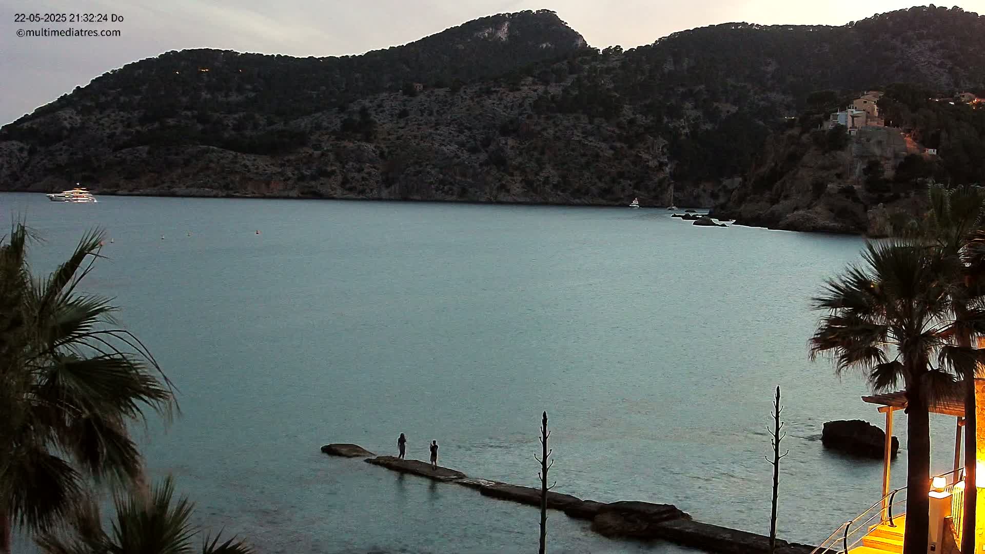Two people stand on a jetty extending into a calm, twilight sea, with a dark, hilly coastline in the background.