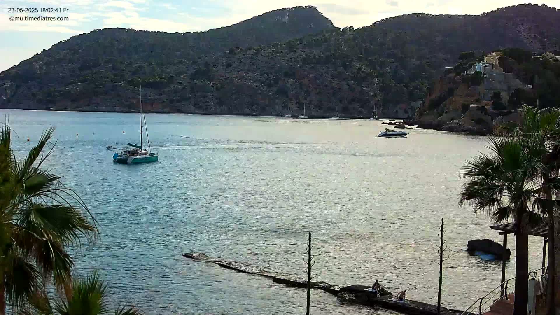 A calm bay with a sailboat and motorboat, surrounded by a hilly landscape with palm trees and a few people near a small jetty under a partly cloudy sky.