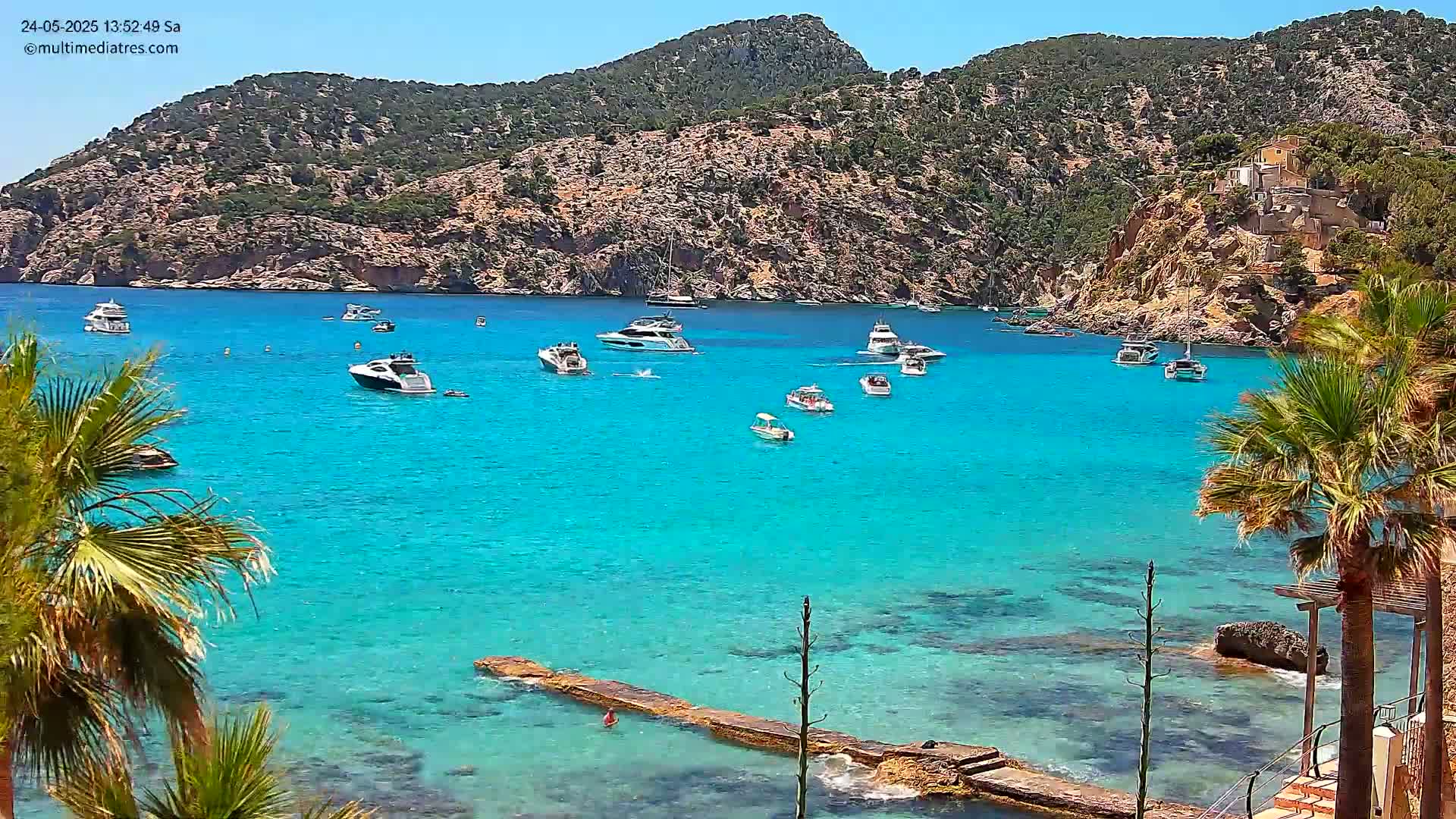 Several boats are anchored in a calm, bright turquoise bay surrounded by rocky hills under a sunny sky.