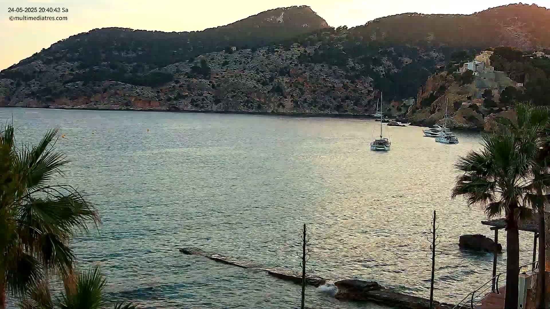 Several sailboats are moored in a calm bay at sunset, surrounded by rocky hillsides with some buildings and palm trees.