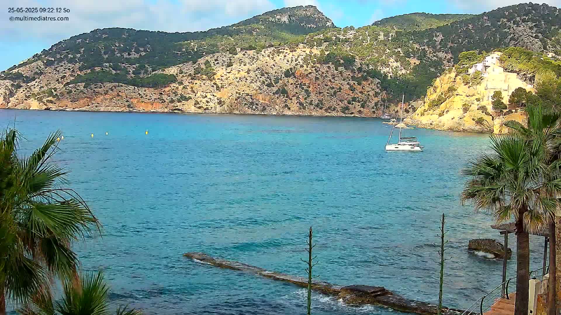 A sailboat is anchored in a calm, turquoise bay surrounded by rocky, green hills under a mostly clear sky.