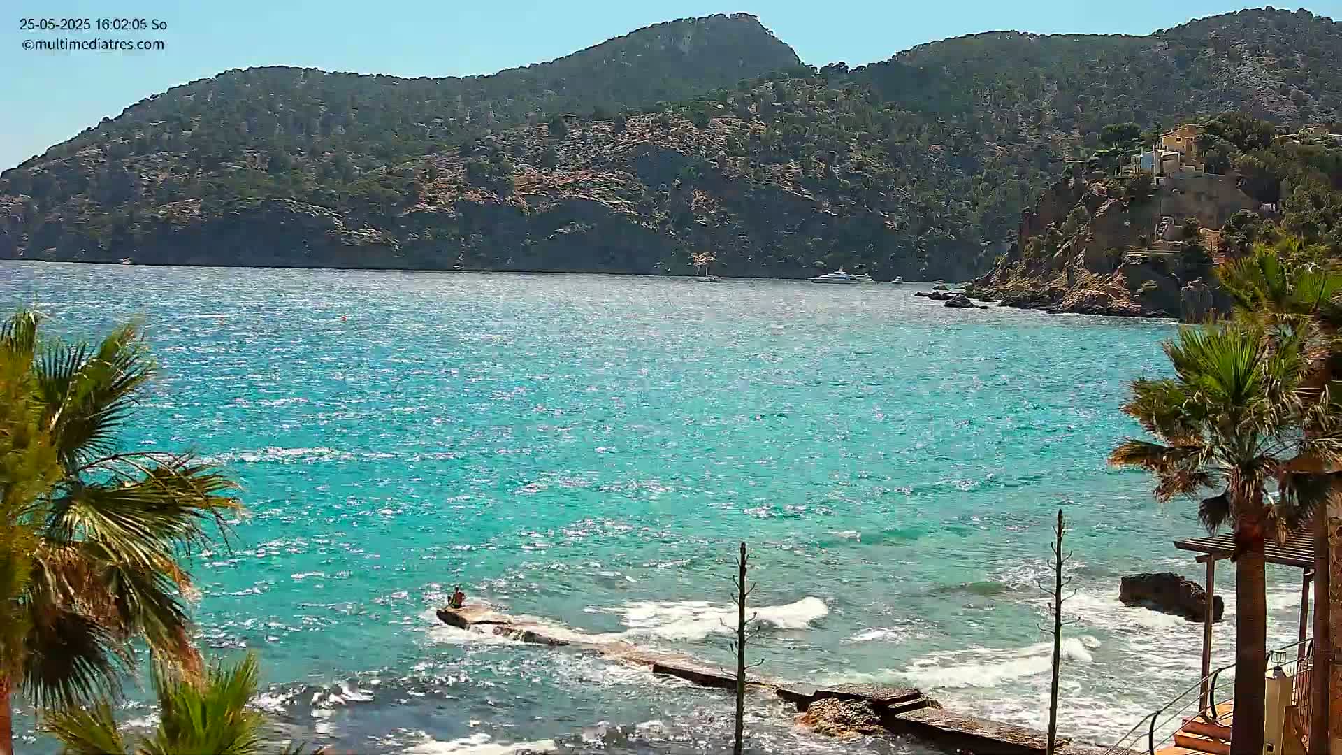 A person sits on a rock jetty extending into a bright turquoise sea lapping against a rocky shore, with a large mountain in the background under sunny skies.