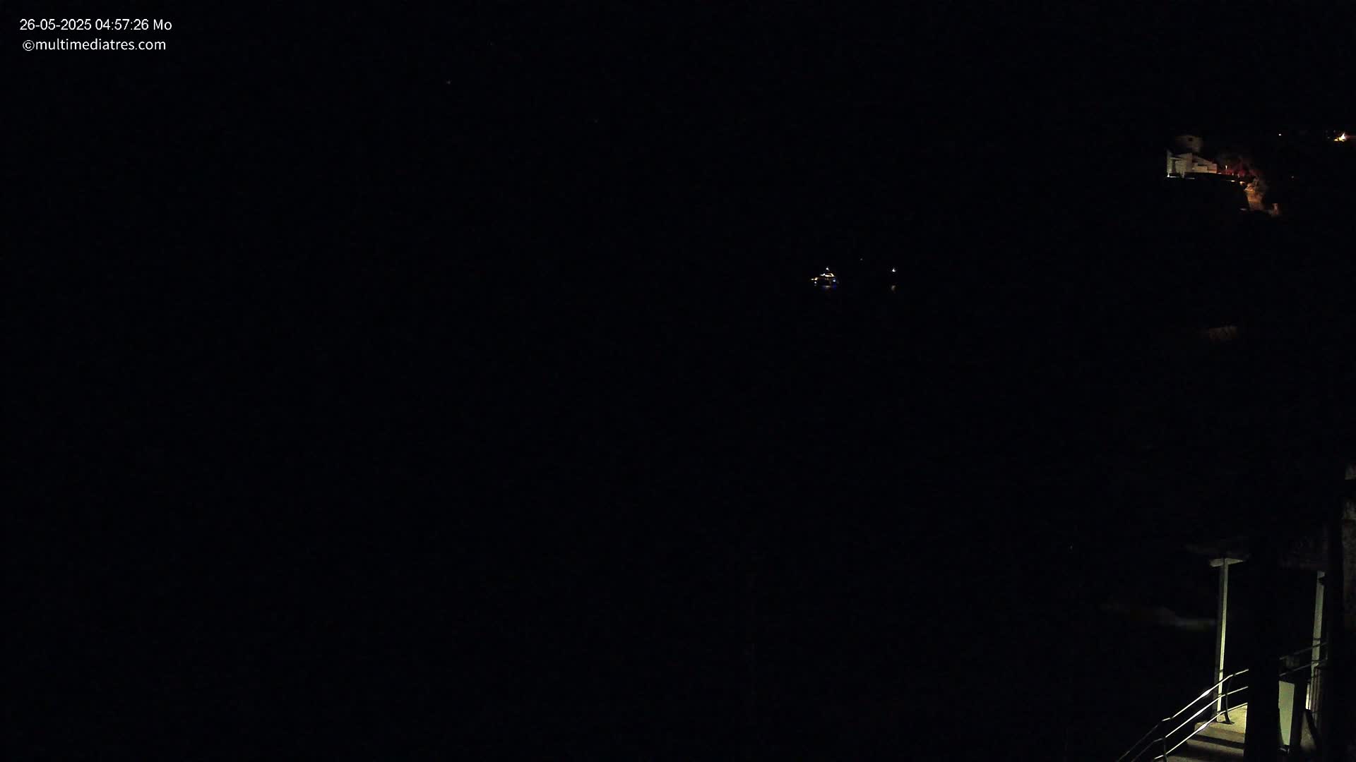 A nighttime view shows a dimly lit structure and faint lights in the distance under a dark sky.