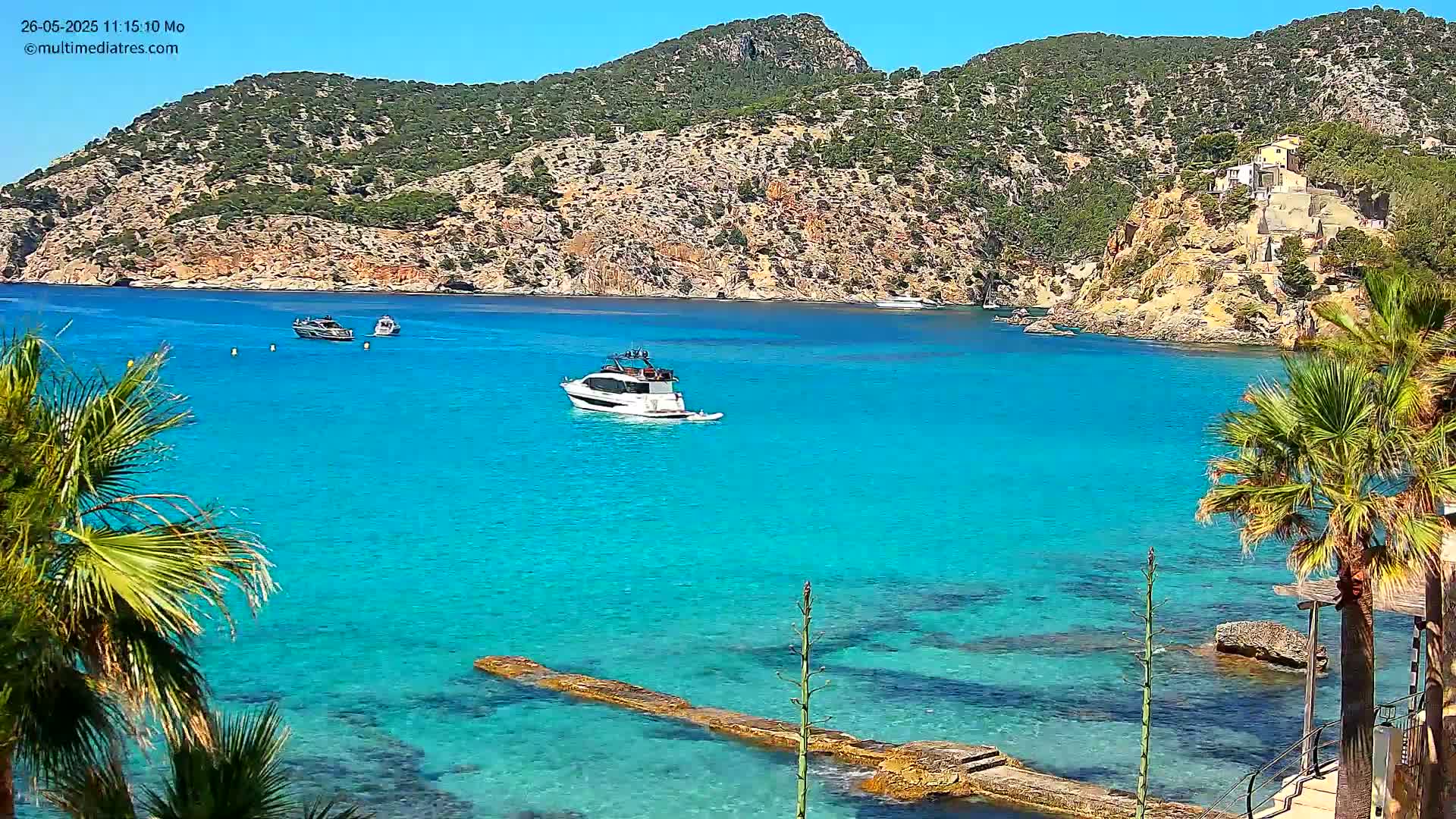 A bright, sunny day reveals a turquoise bay with several boats, partially visible palm trees, and a rocky hillside.
