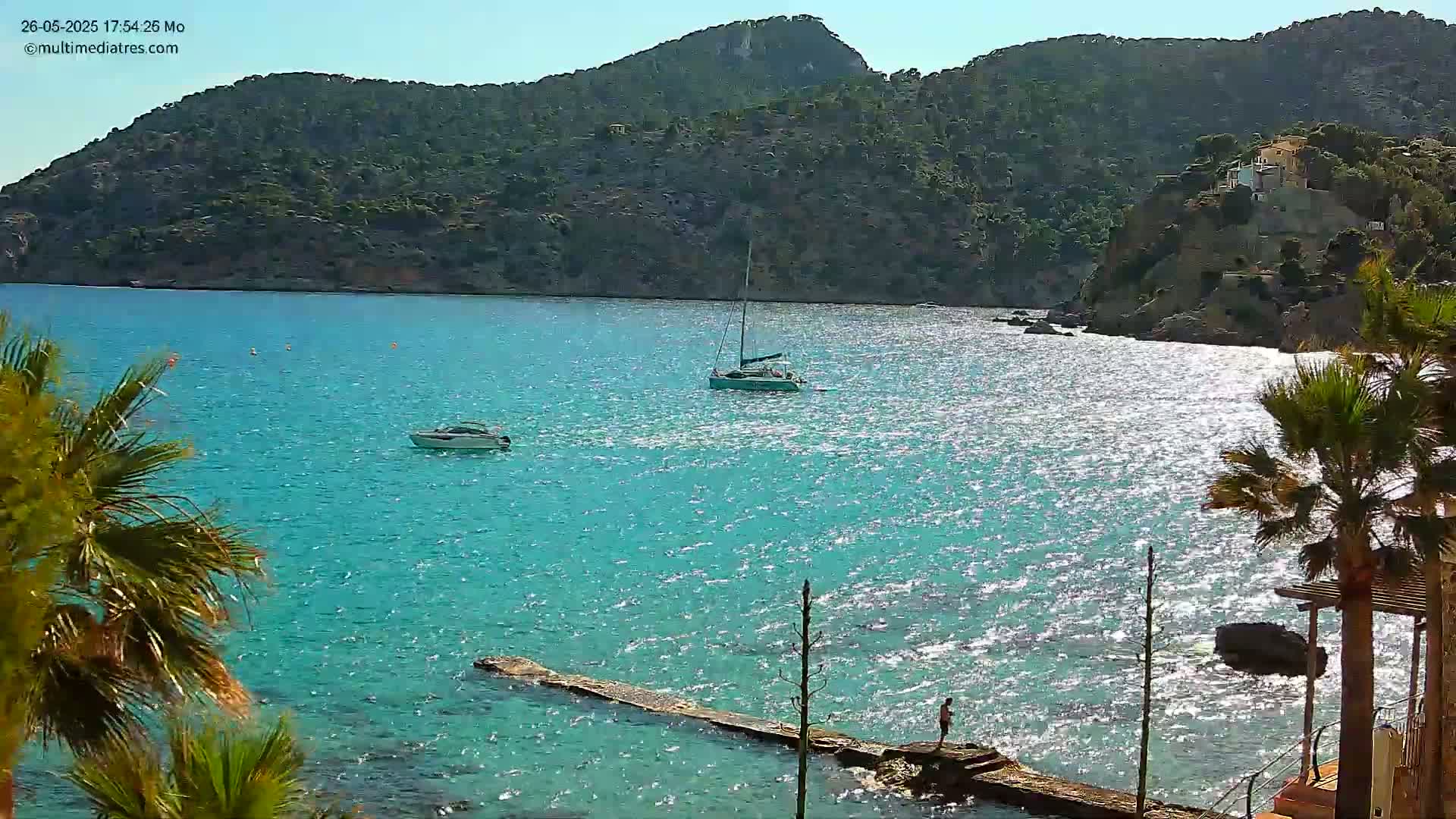 A sunny day shows a bright turquoise bay with several small boats, a long wooden pier, and palm trees on a rocky shore beneath a green hill.
