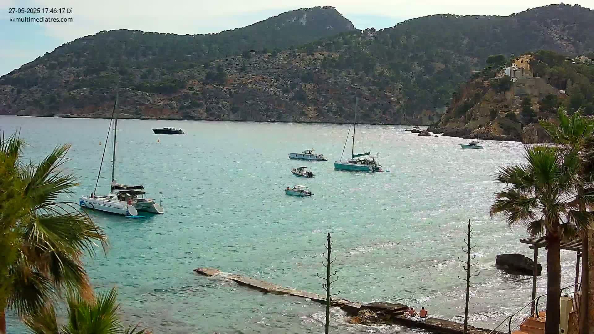 Several boats are anchored in a calm, turquoise bay surrounded by rocky, green hills under a mostly clear sky.