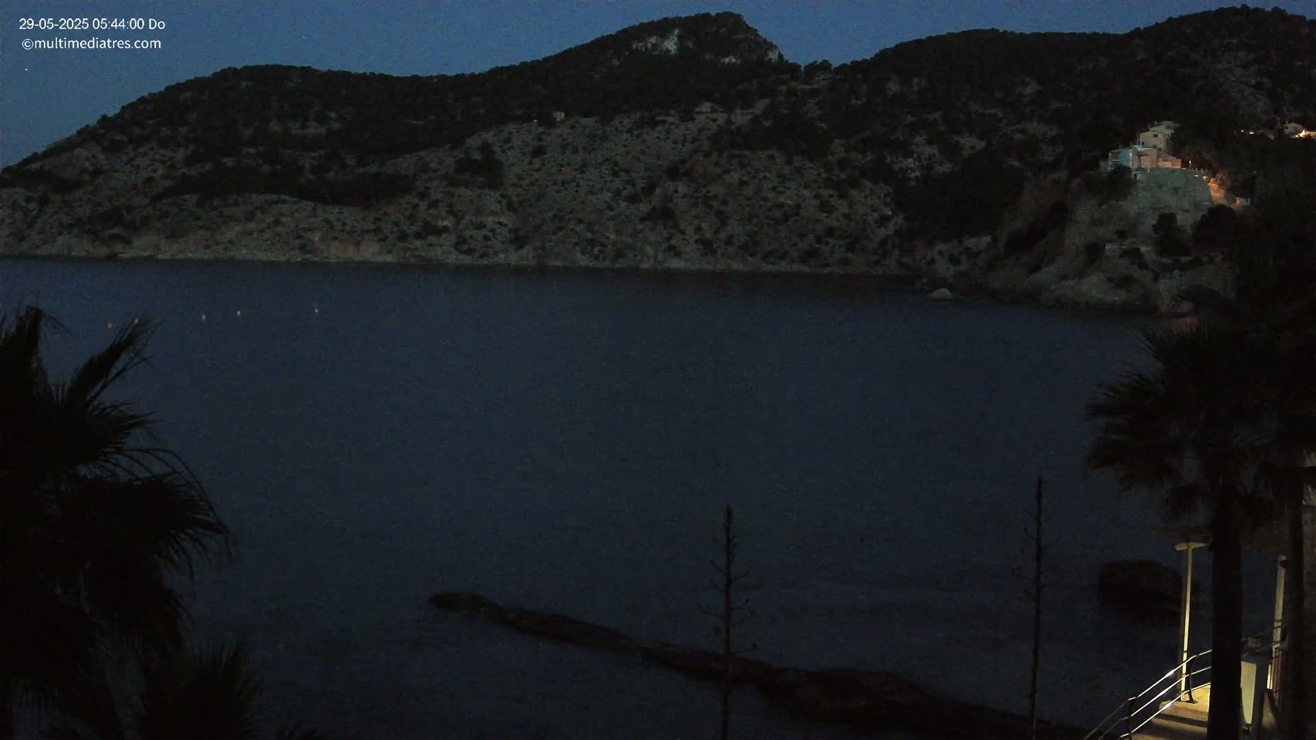 A dark, still body of water is seen at twilight, with a mountainous shoreline silhouetted against the darkening sky, partially obscured by palm trees in the foreground.