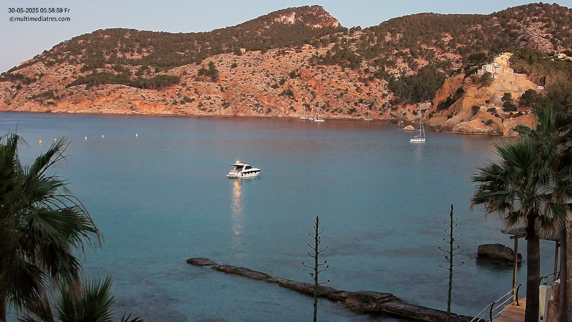 A calm, blue bay is nestled between reddish-brown hills dotted with vegetation, a single motorboat sits in the water near a rocky shoreline partially obscured by palm trees.