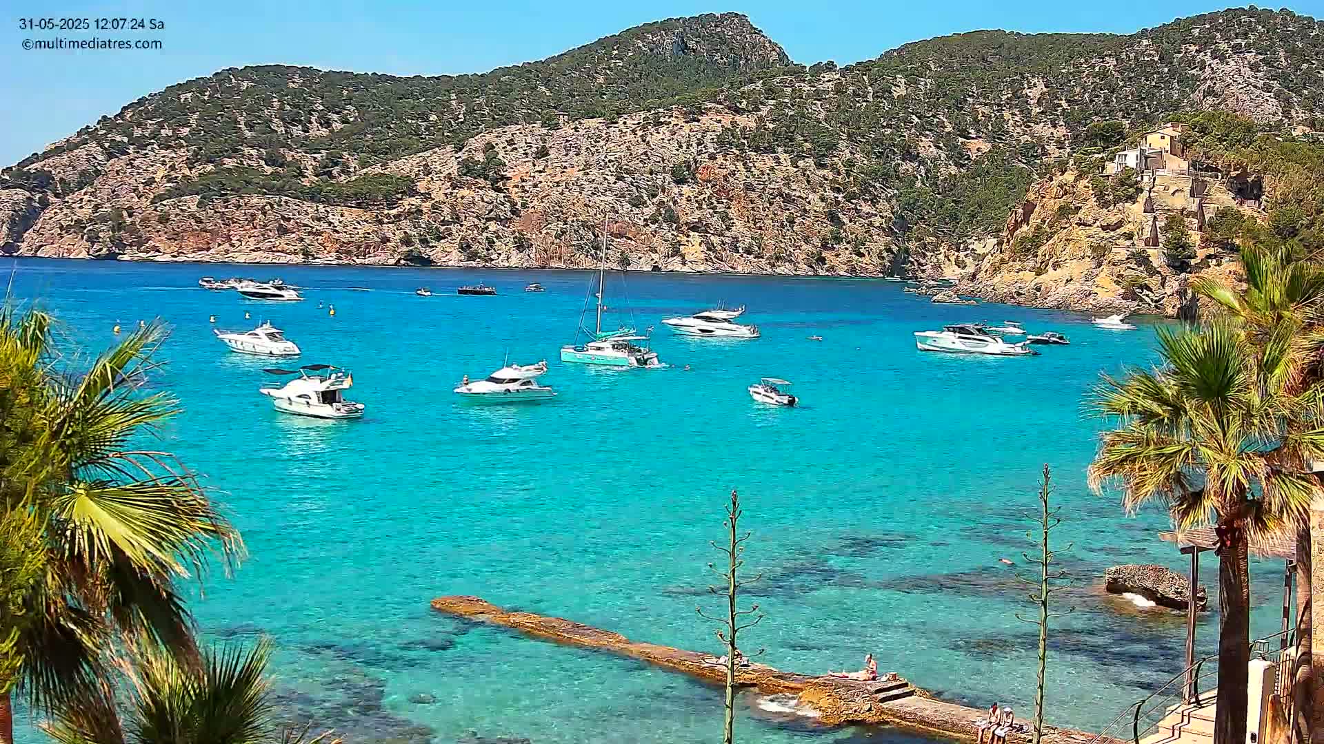 Several boats are anchored in a clear, turquoise bay surrounded by a rocky, green hillside on a sunny day.