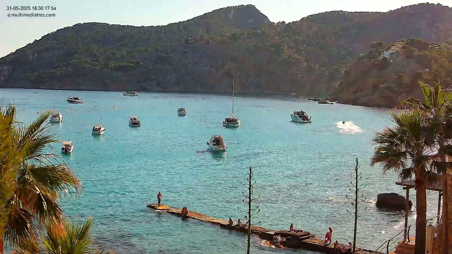 A calm, clear blue bay with numerous boats and a few people on a jetty, surrounded by a rocky hillside under a sunny sky.