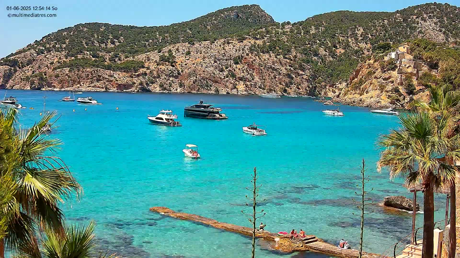 A sunny day shows a calm, bright turquoise bay with several boats anchored, surrounded by rocky hillsides dotted with vegetation and a few people relaxing on a low stone jetty.