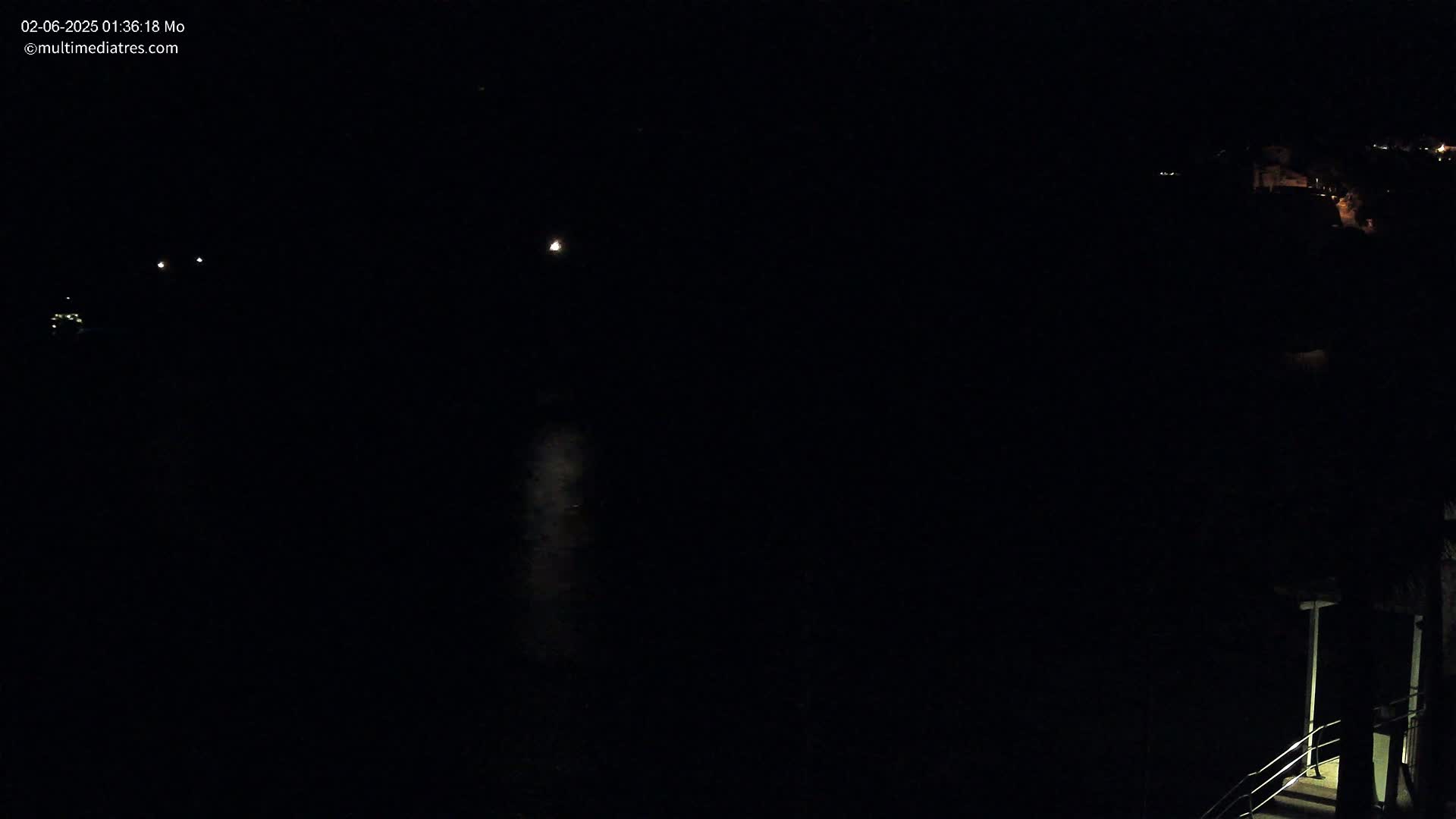 A dark nighttime scene shows a body of water reflecting faint lights, with dimly lit structures visible in the distance and a stairway on the lower right.